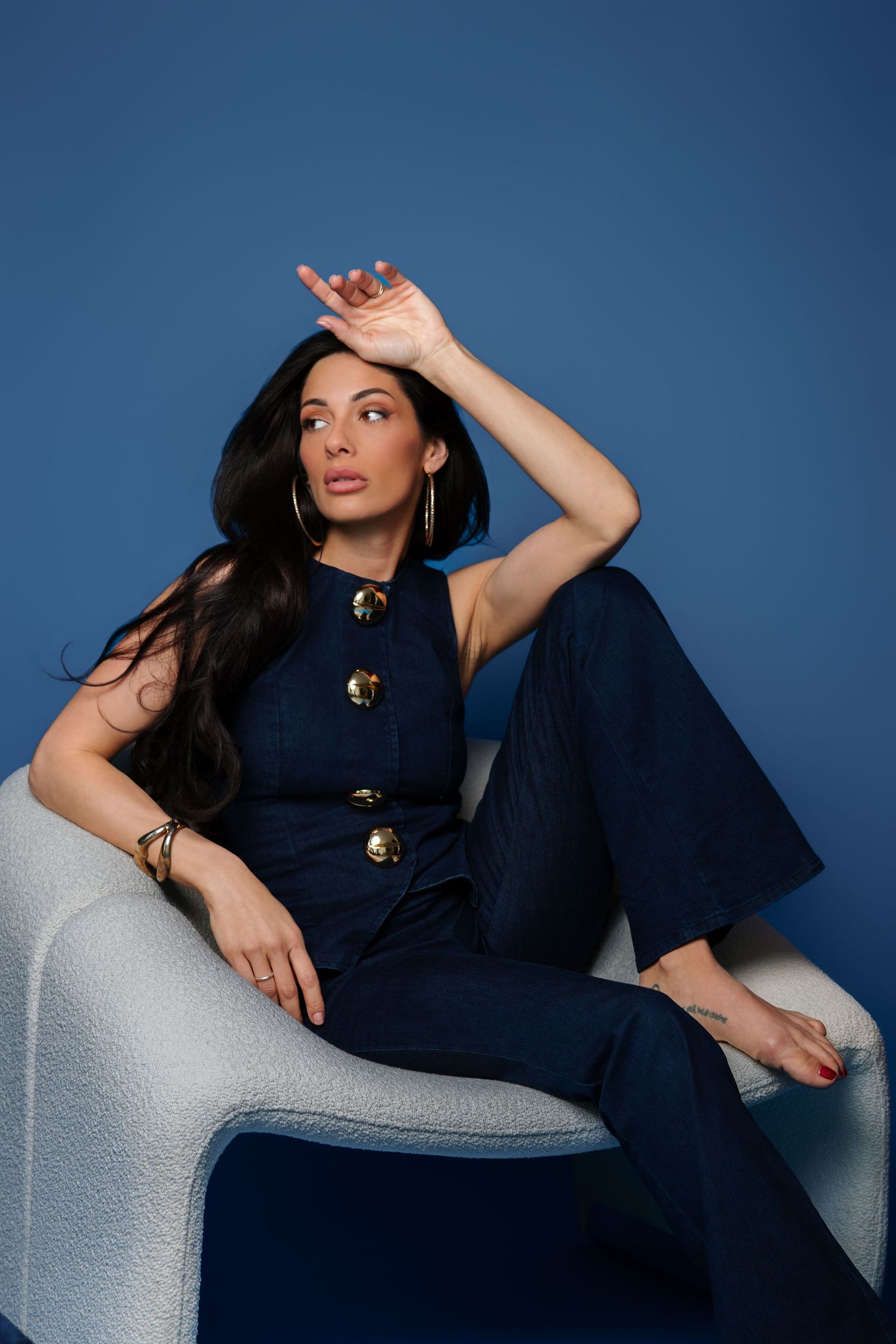Stylish dark-haired woman in navy jumpsuit sitting in modern chair against vibrant blue studio background.
