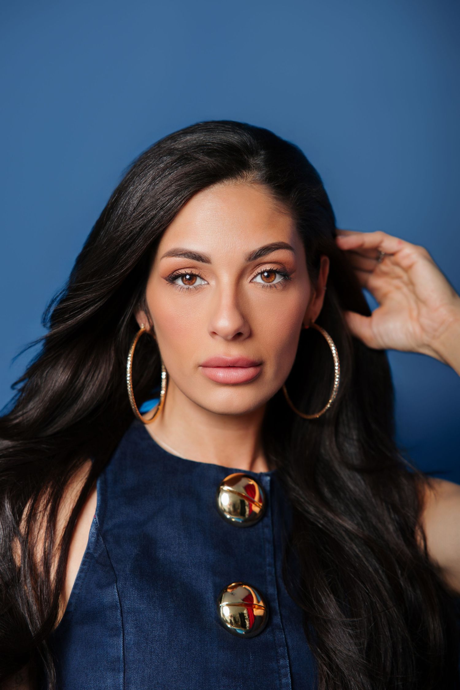 Beautiful woman with long dark hair wearing gold hoop earrings and denim top against blue backdrop.