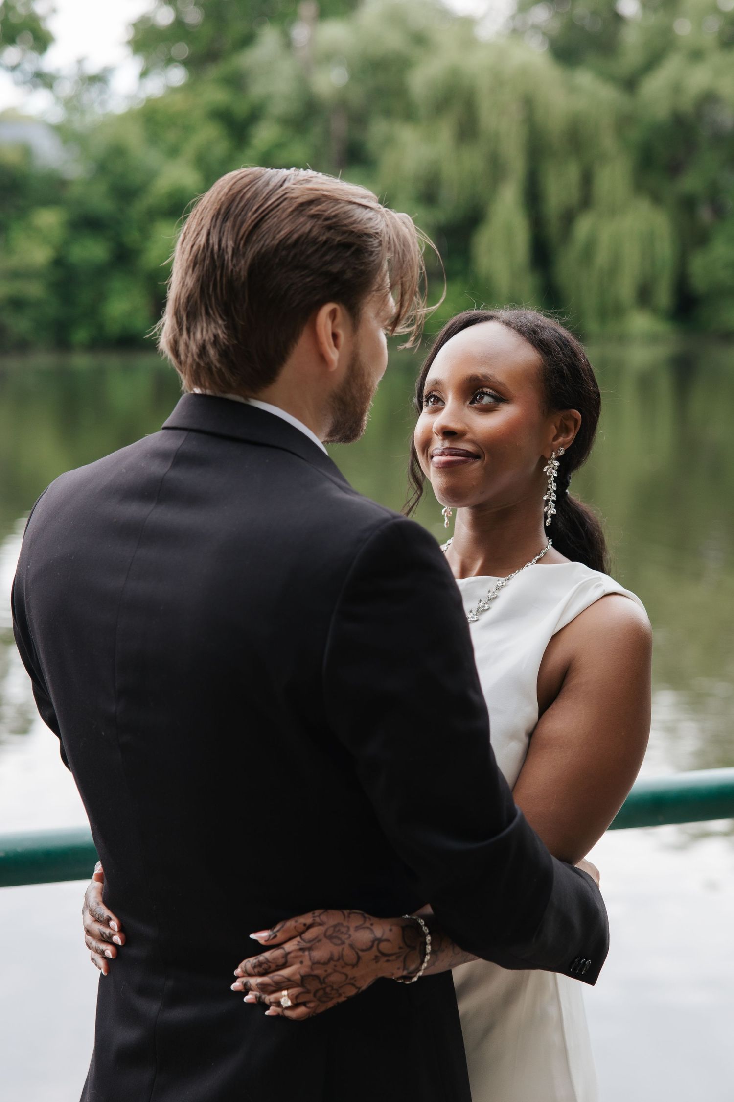 Interracial couple embracing near a pond, bride in white dress smiling at groom in black suit outdoors.