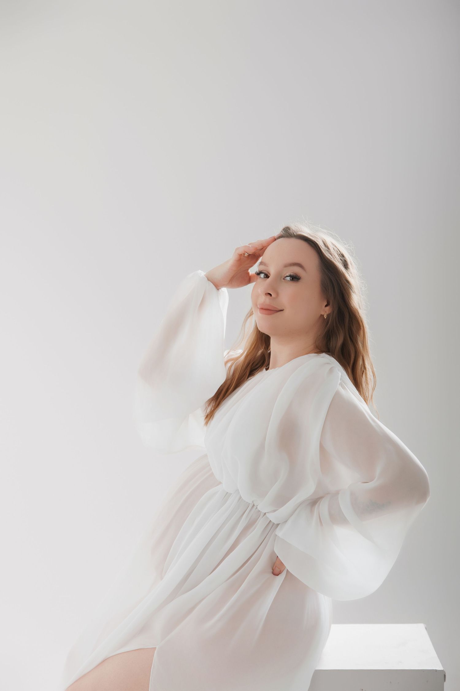 Young woman in white chiffon dress posing gracefully against a light gray background in a studio setting.