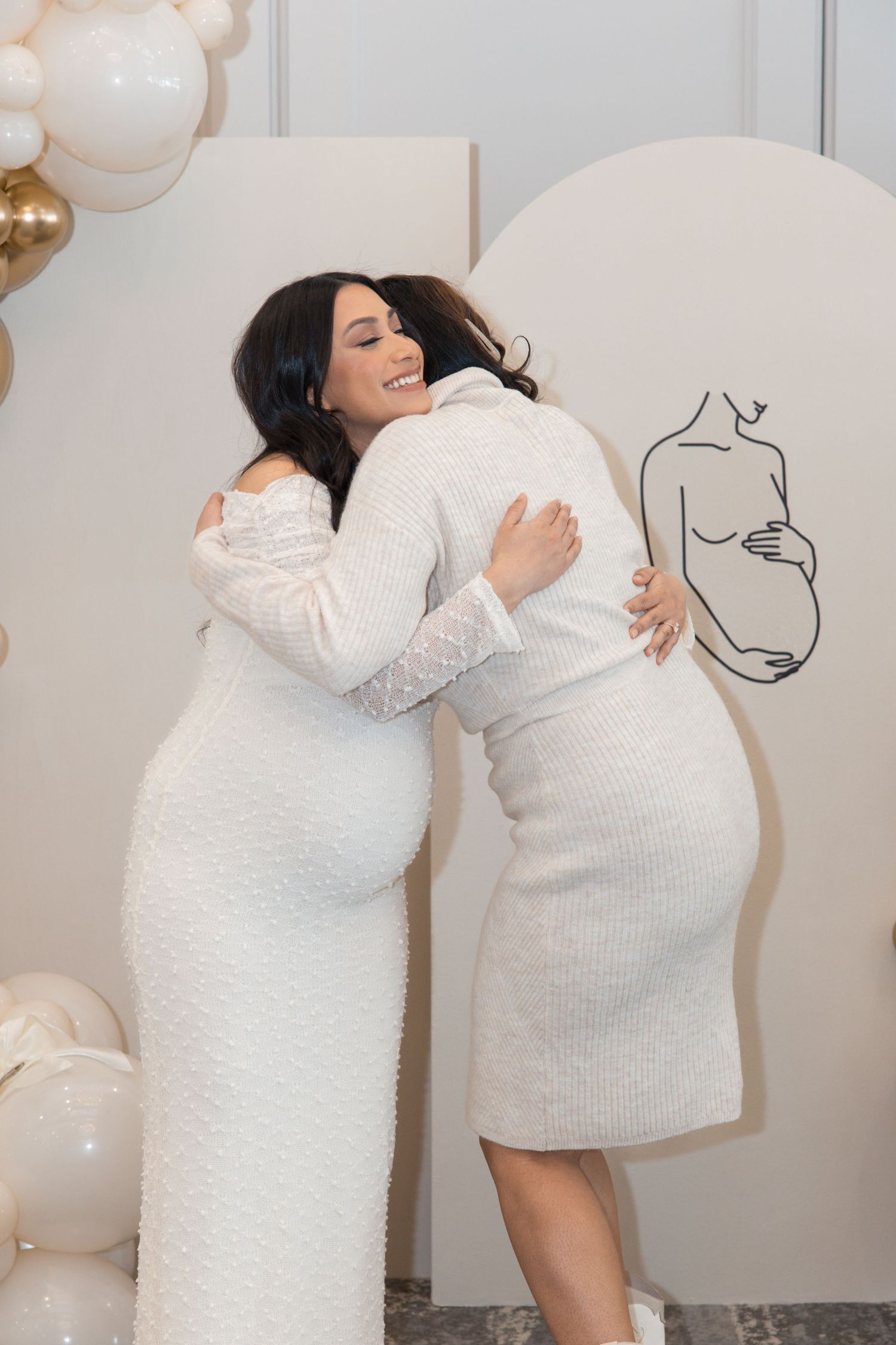 Two pregnant women in white dresses hugging and smiling at a baby shower with balloon decorations.