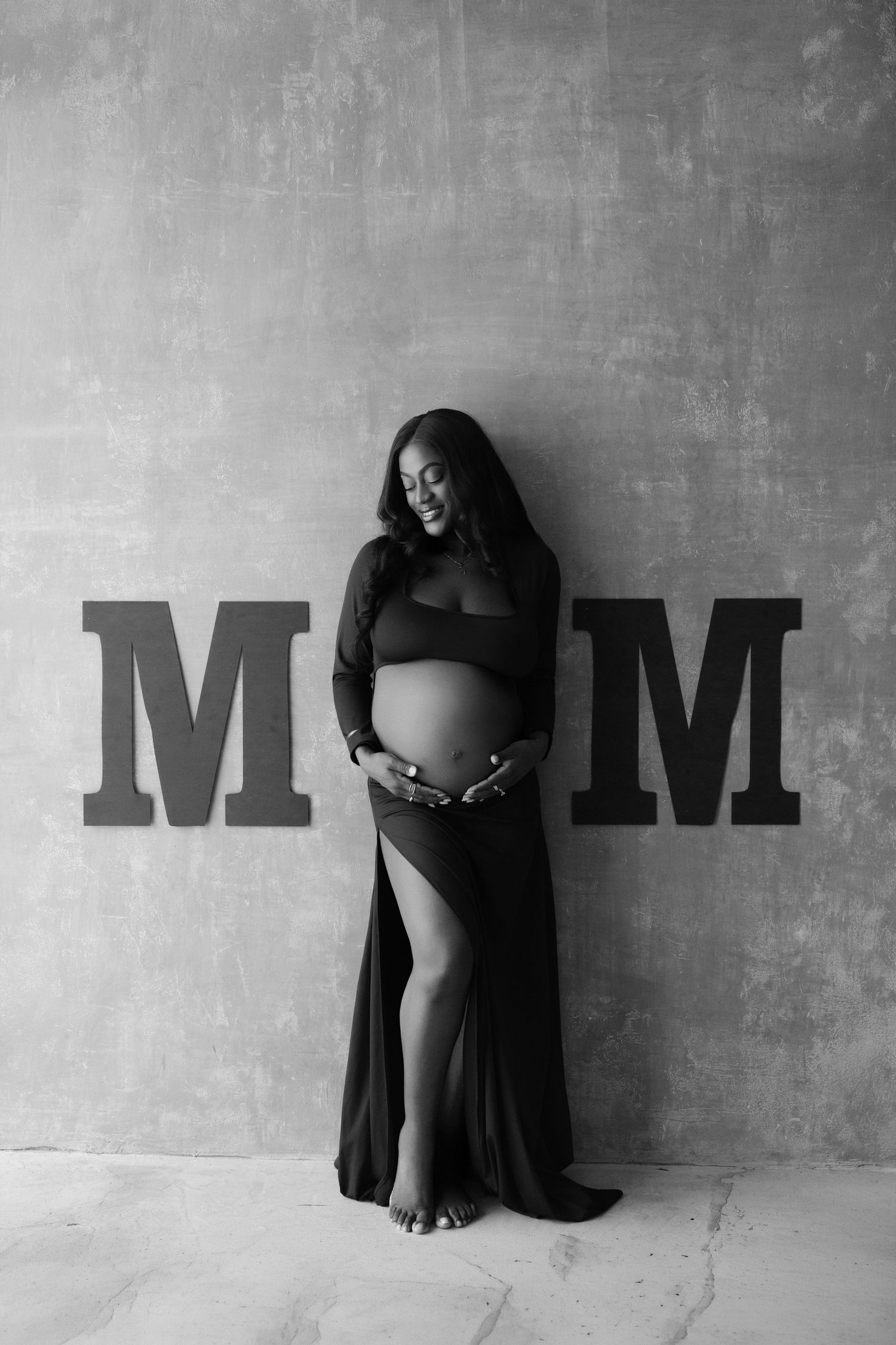 Black and white maternity photo of pregnant woman in black dress posing between MOM letters against gray wall.