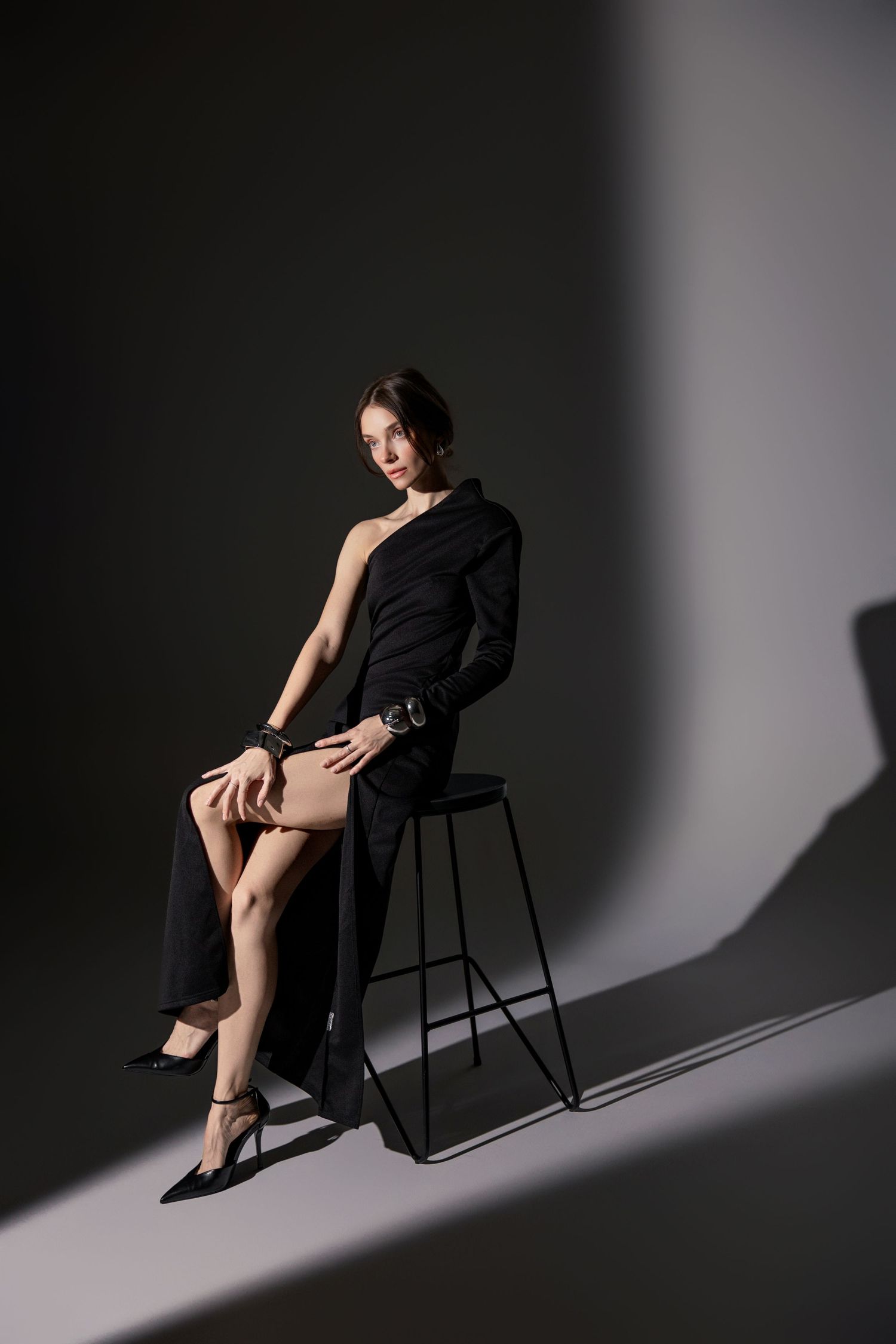 Woman in asymmetric black blazer dress seated on stool in dark studio.