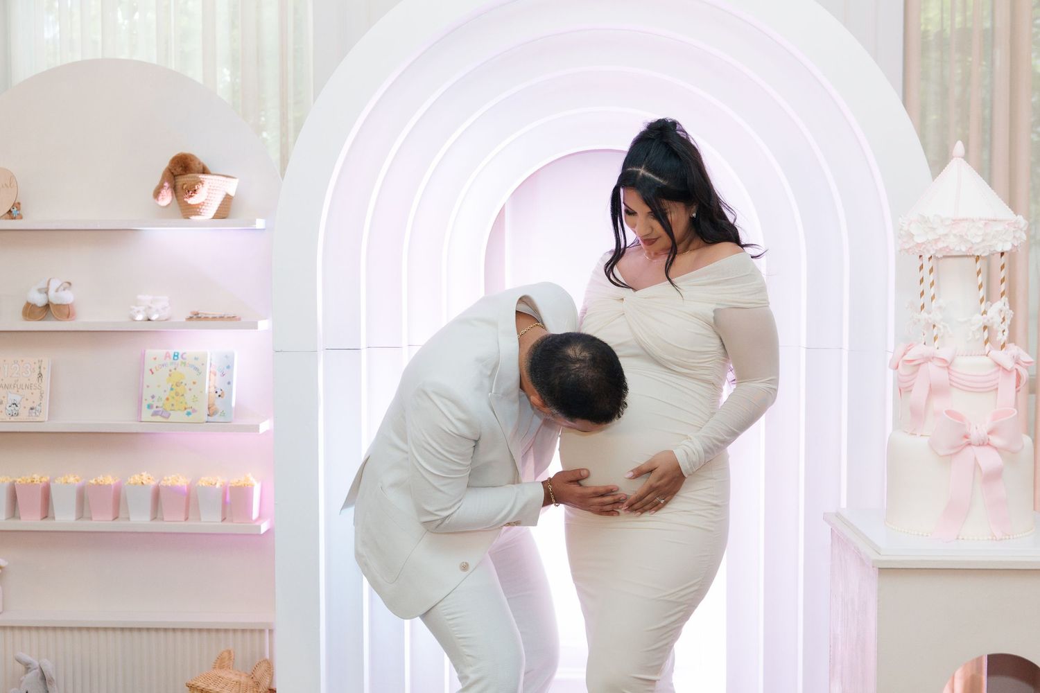 A man in white kneels to kiss a pregnant woman's belly during an elegant baby shower celebration.