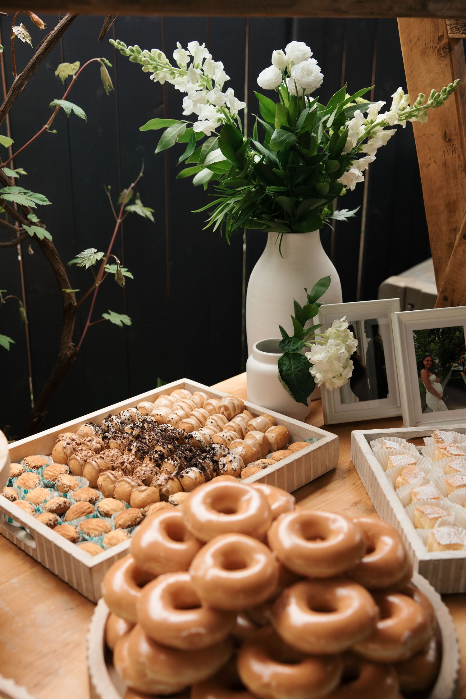 Elegant dessert table with glazed donuts, assorted pastries, and white floral arrangement in a vase at a wedding reception.