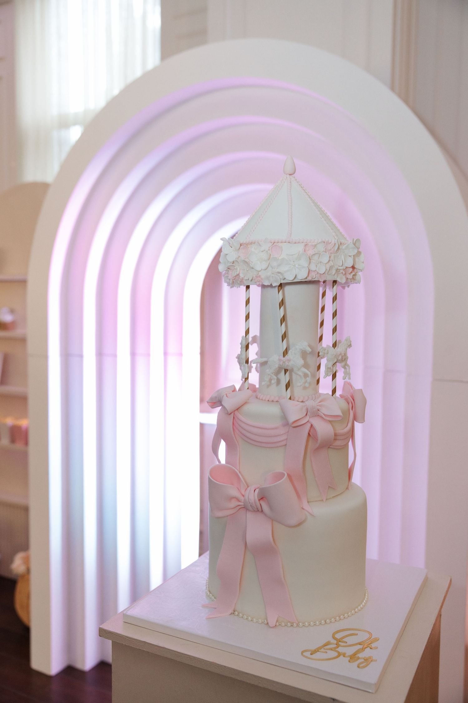 Elegant white carousel cake with pink bows and gold accents displayed under a glowing pink arch backdrop.