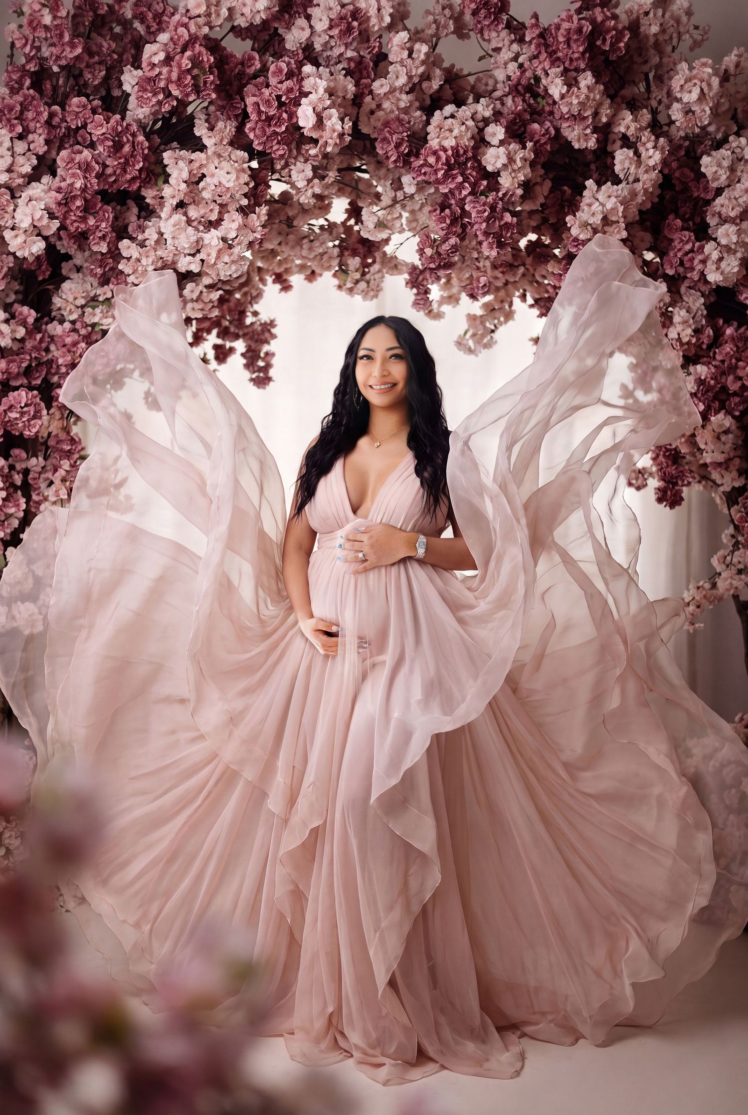 Pregnant woman in flowing pink gown poses gracefully under floral arch with mauve blooms in elegant maternity photo.