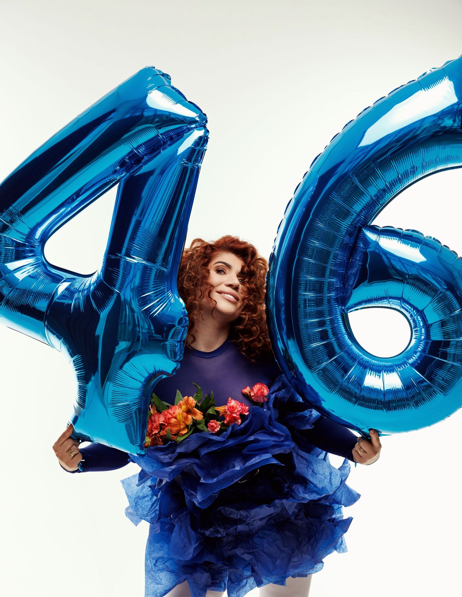 Smiling curly redhead woman in blue ruffled dress holding oversized blue number 46 balloons and colorful flowers indoors.