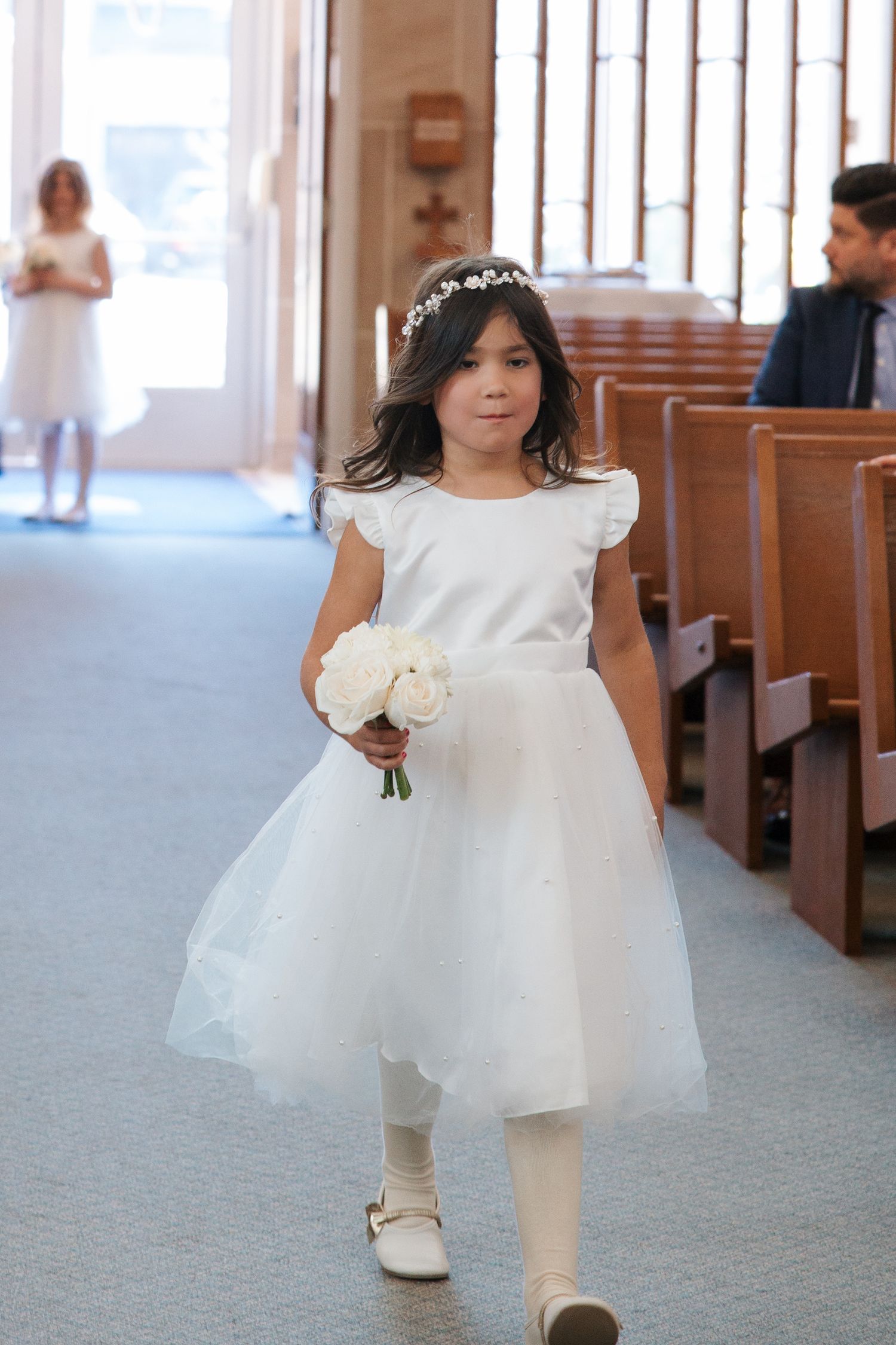 Young flower girl in white tulle dress and floral crown walks down church aisle holding white rose bouquet.
