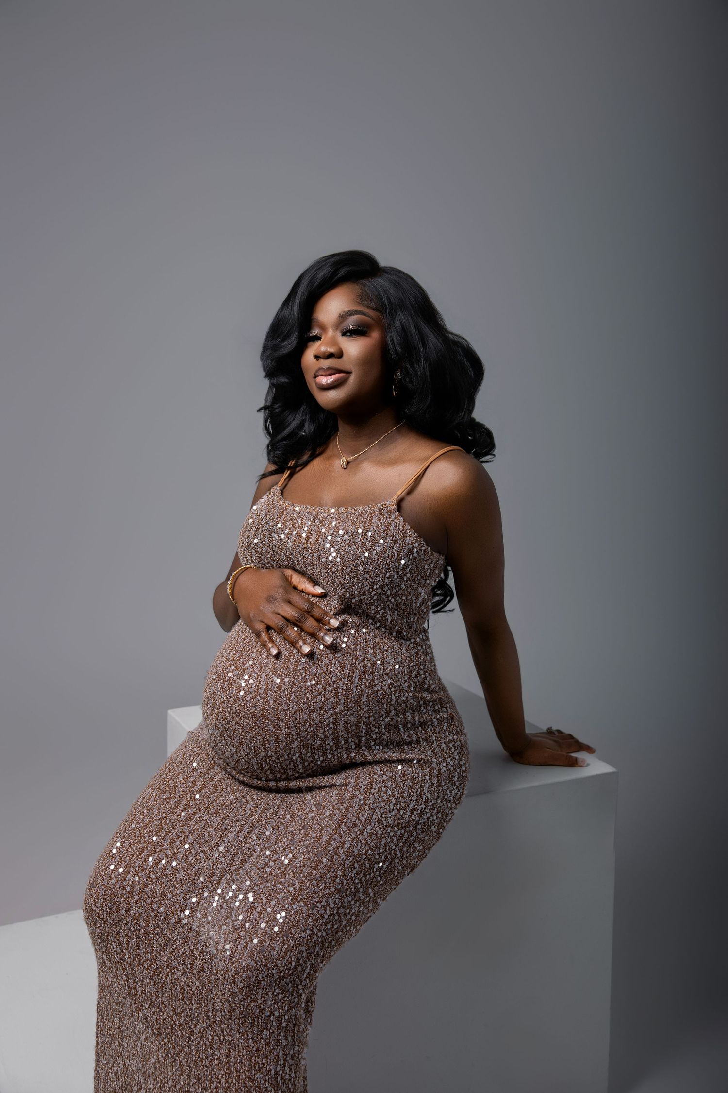 Pregnant woman in a sequined rose gold gown cradling her baby bump, posing elegantly against a gray studio background.