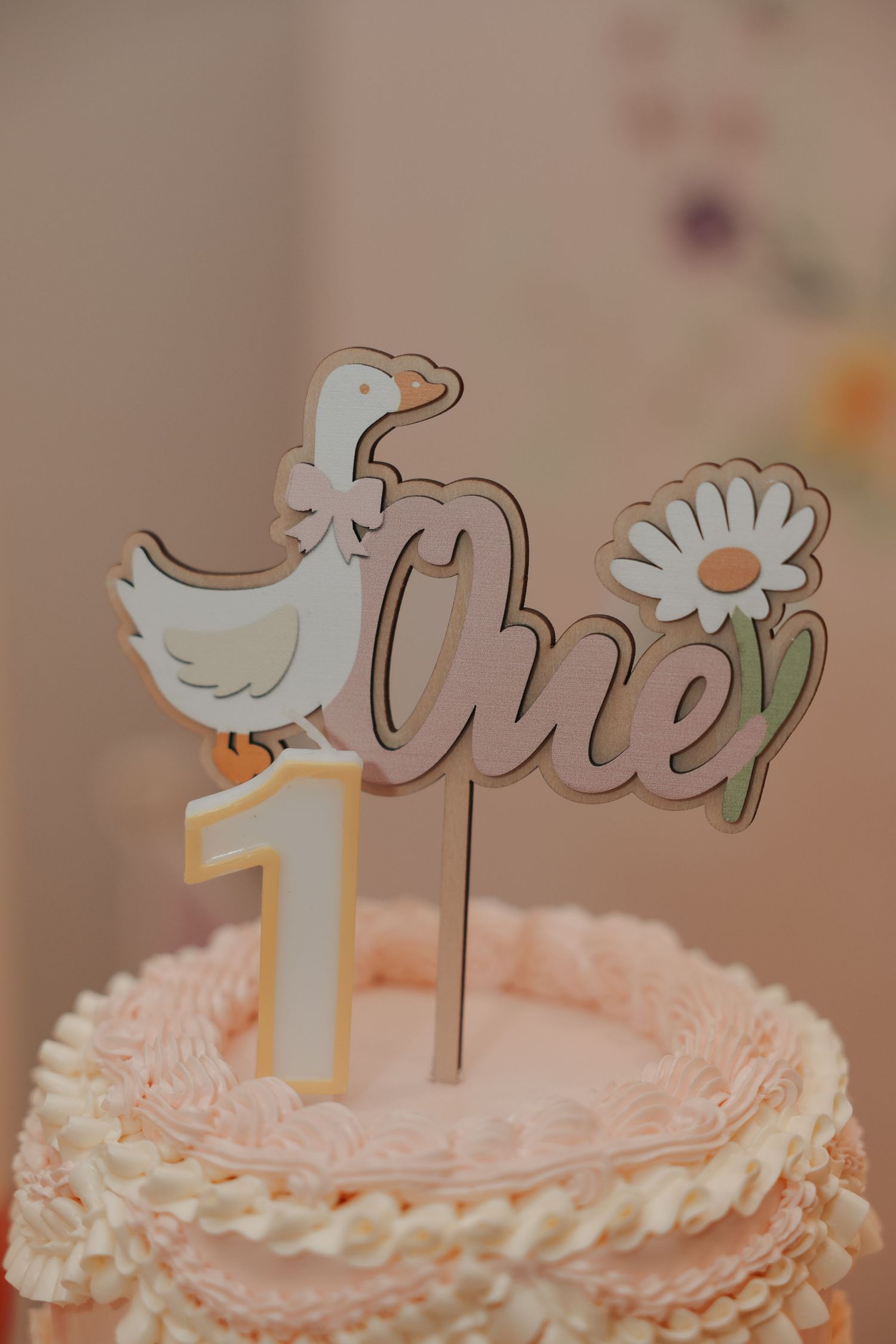 Pink first birthday cake with duck and daisy topper reading One, decorated with ruffled frosting.