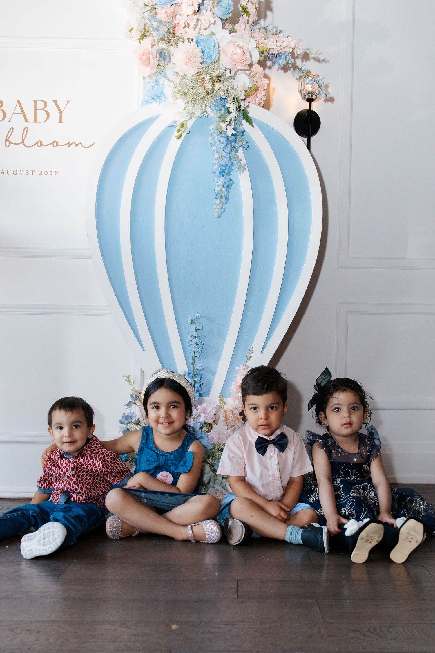 Four young children sitting on floor in front of Baby Bloom party backdrop with hot air balloon decoration.