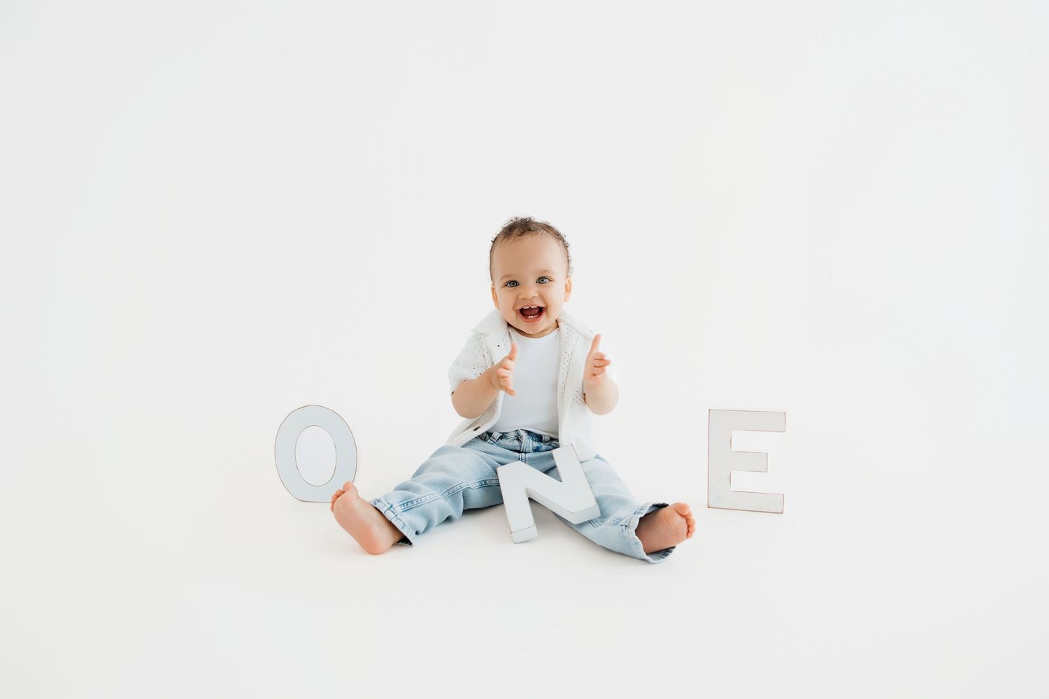 A happy baby boy sits on a white background wearing jeans, surrounded by letters spelling ONE for his first birthday.