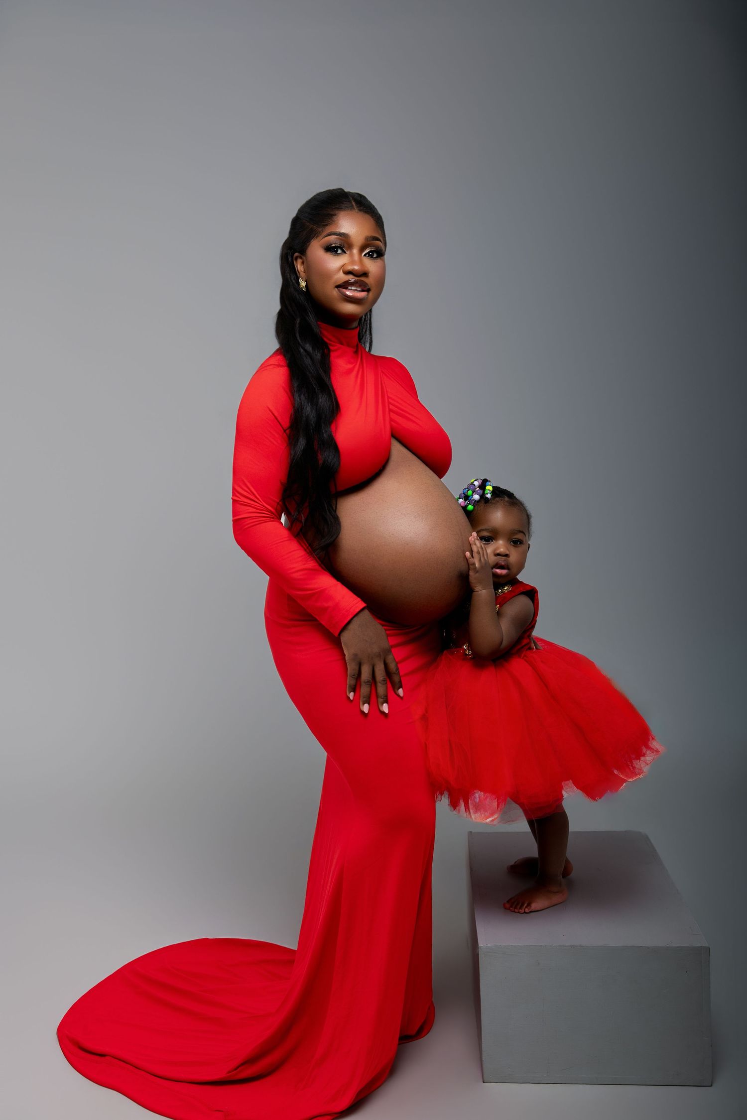 Pregnant Black woman in red maternity gown poses with toddler daughter in matching red tutu dress in studio.