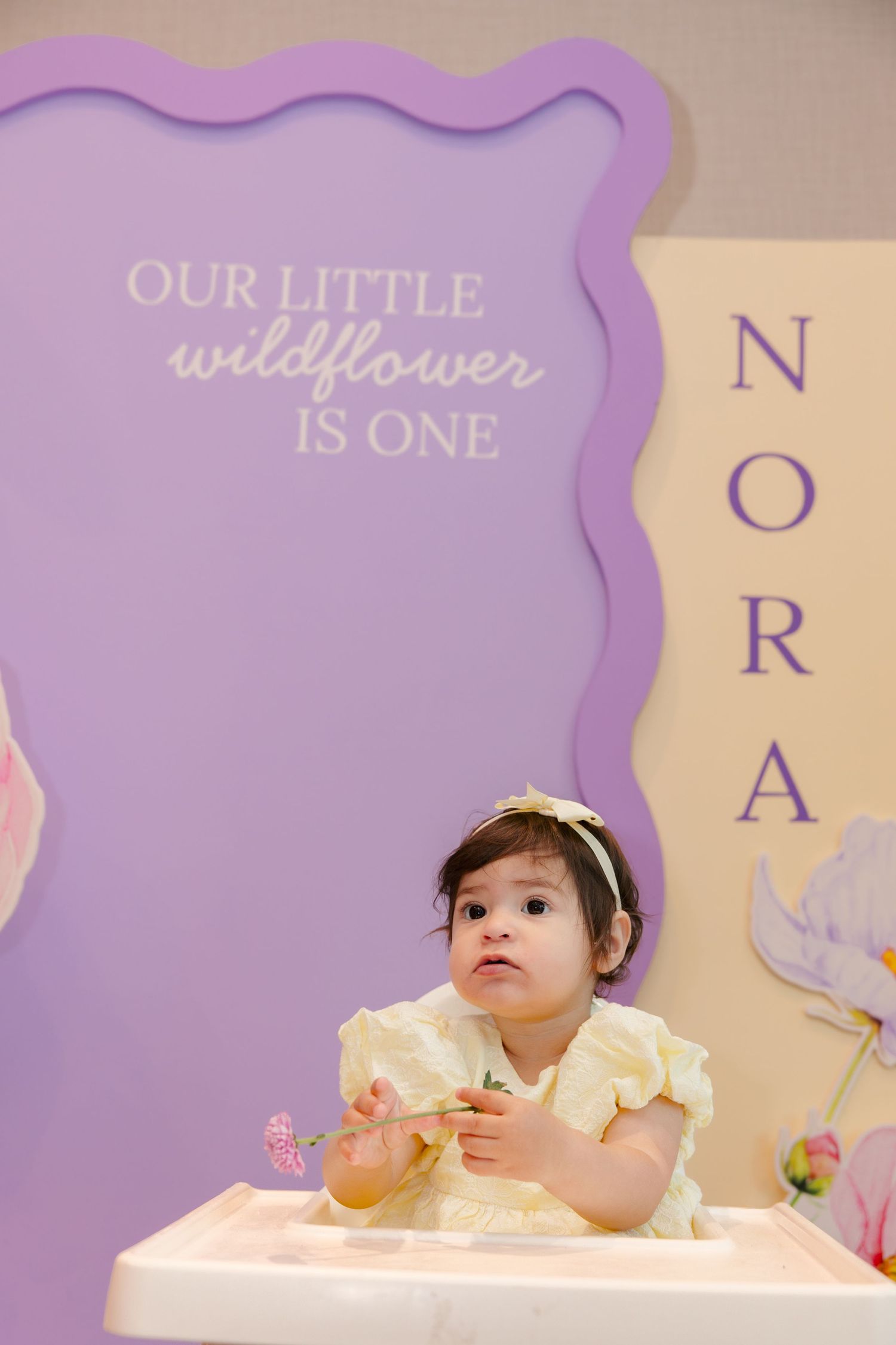 Baby girl in yellow dress sits in high chair at her first birthday party with purple wildflower themed backdrop.