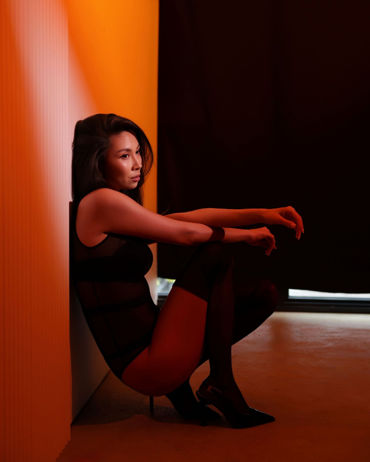 Woman in black outfit sitting against orange wall in dramatic lighting.