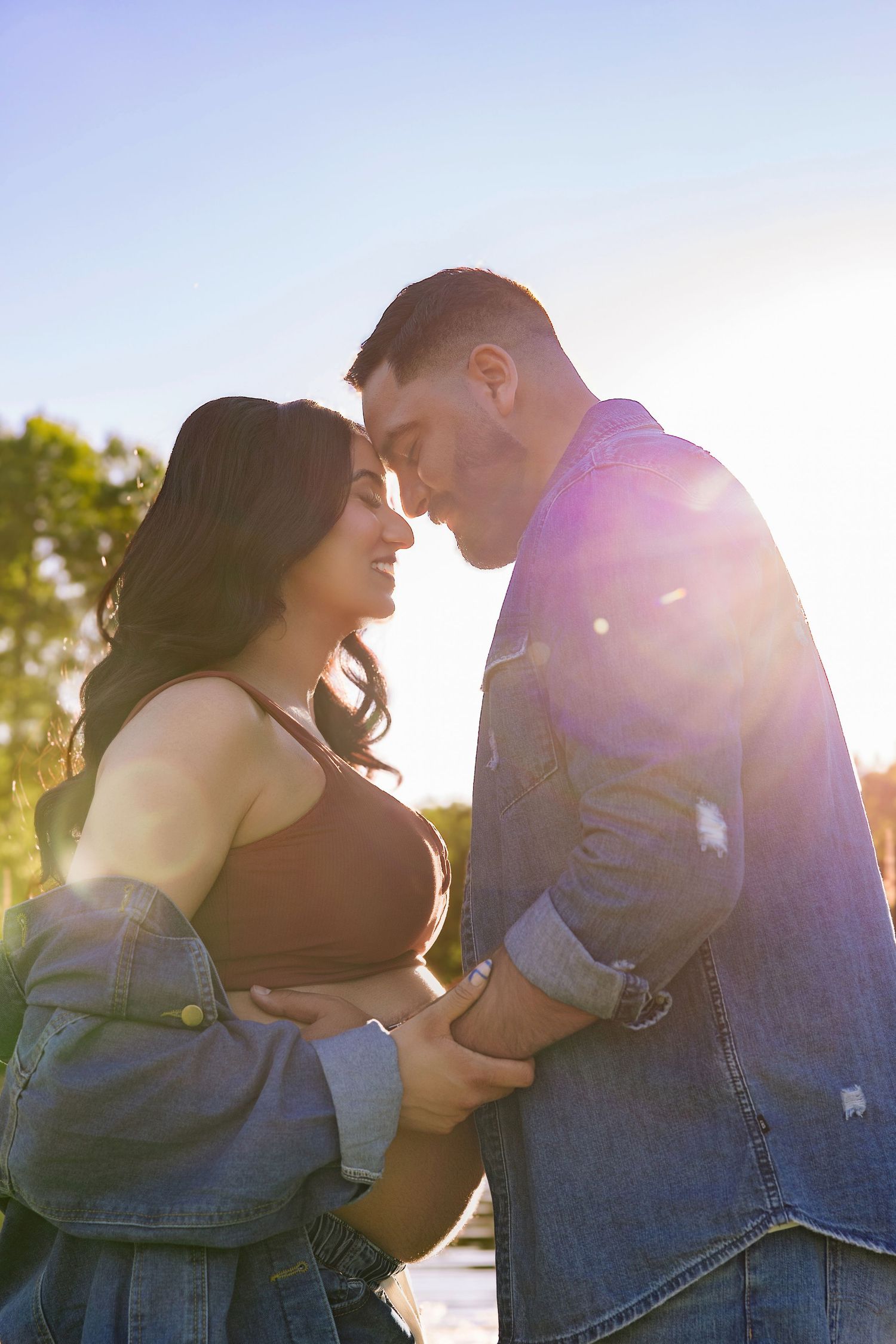 Couple sharing a romantic moment outdoors, bathed in golden sunlight, wearing denim jackets near a serene lake.