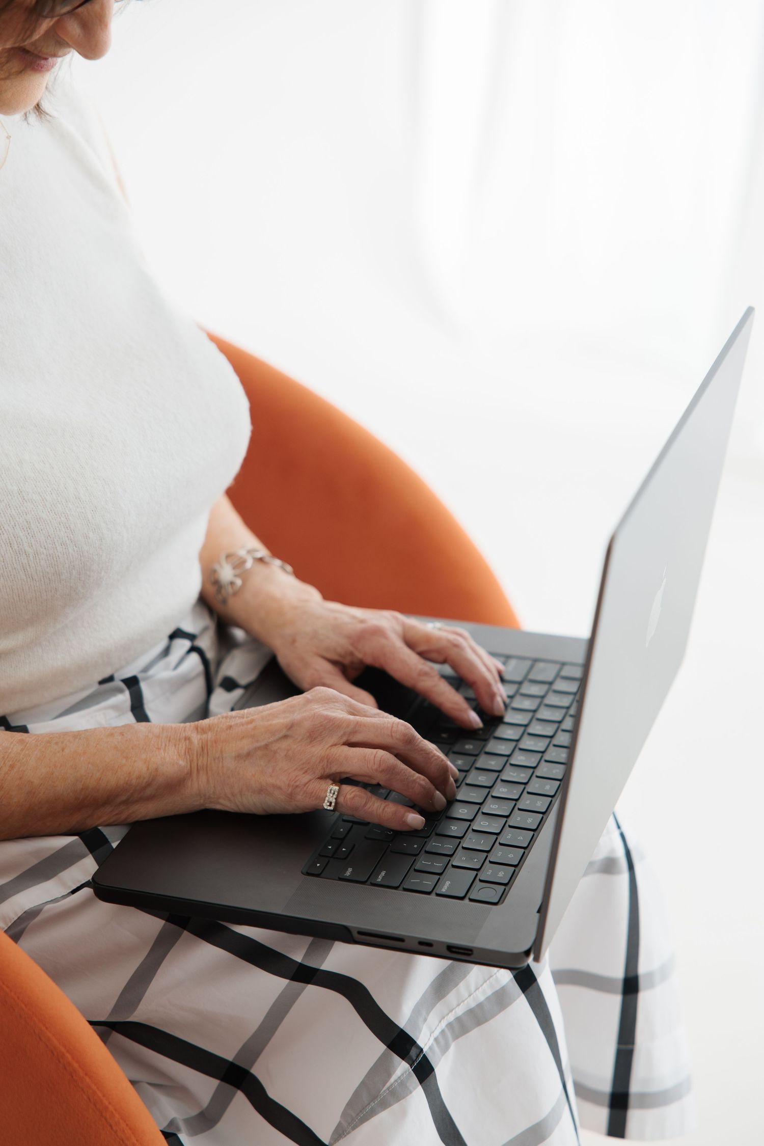Close-up of a woman's hands typing on a laptop while seated in an orange chair.