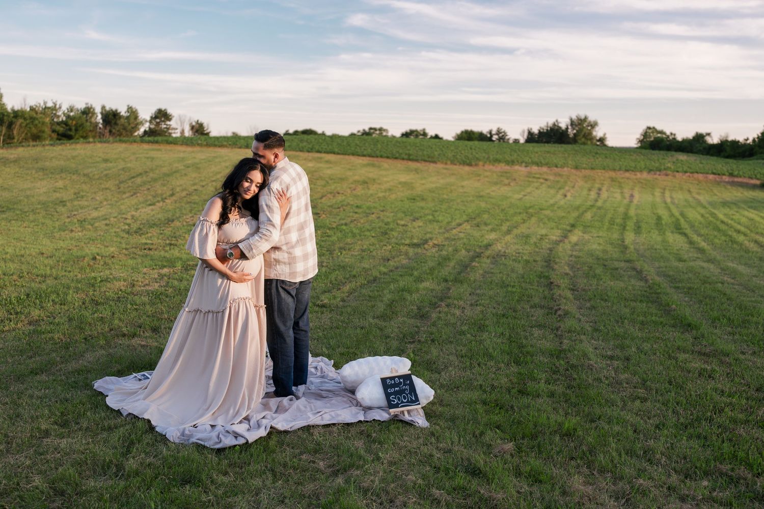 Couple embracing in a green field during a maternity photo session at golden hour with a blanket on the grass.