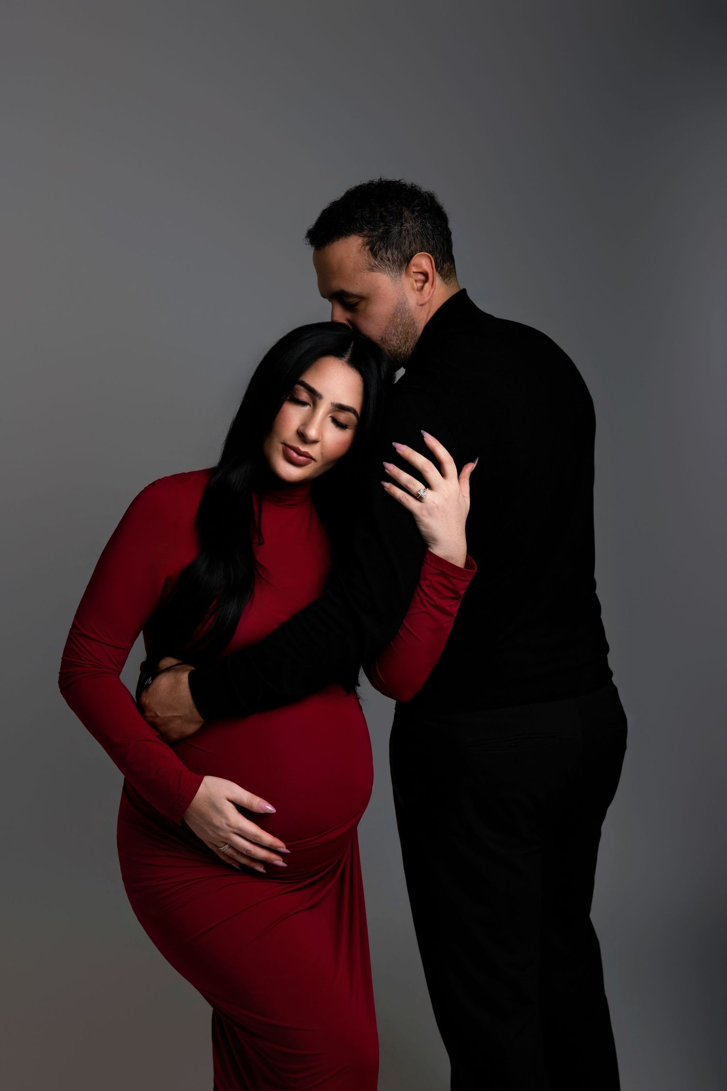 Pregnant woman in red dress with partner in black embracing her from behind in a studio maternity photo.