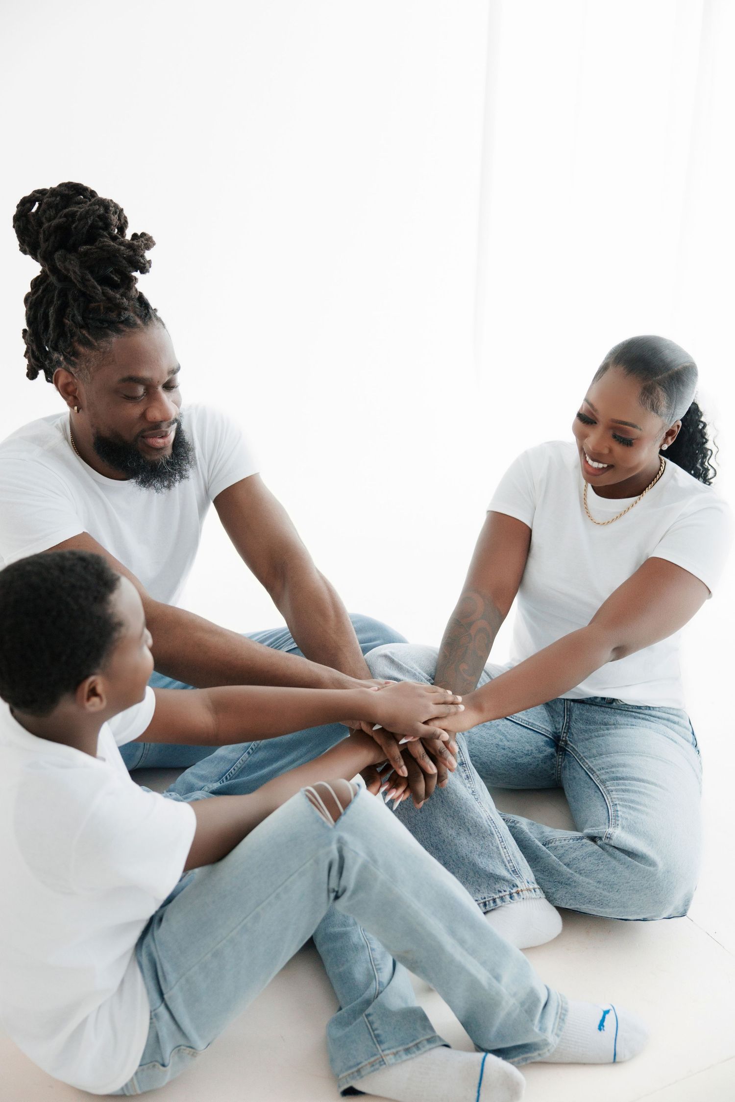 African American family in white tees and jeans sitting together, stacking hands in unity gesture on white background.