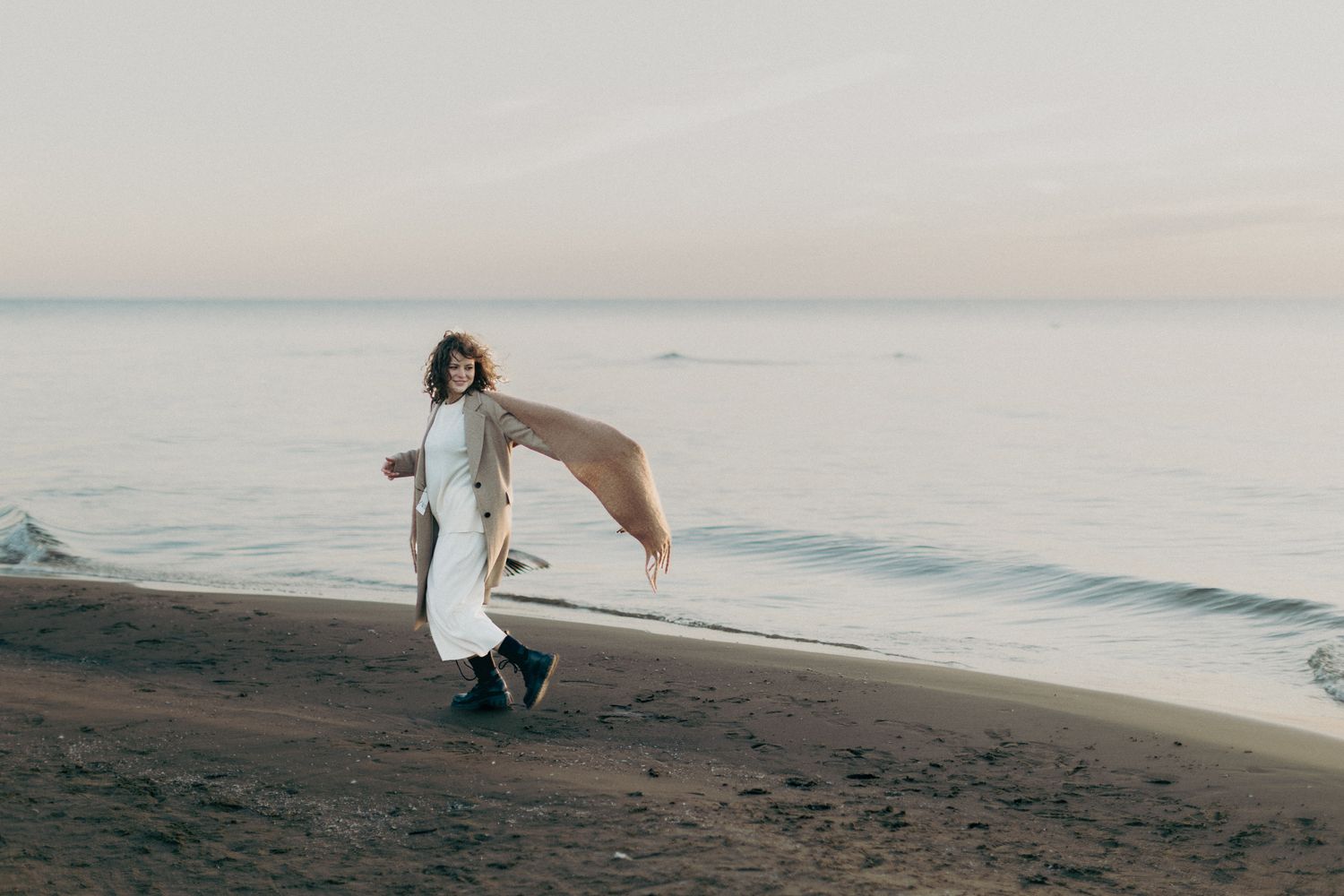 Woman in white dress with flowing scarf walking along beach at sunset.