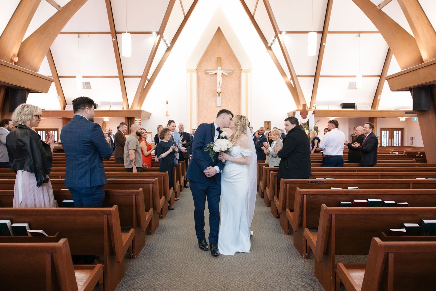 Newlywed couple kissing in church aisle surrounded by applauding guests during wedding ceremony.