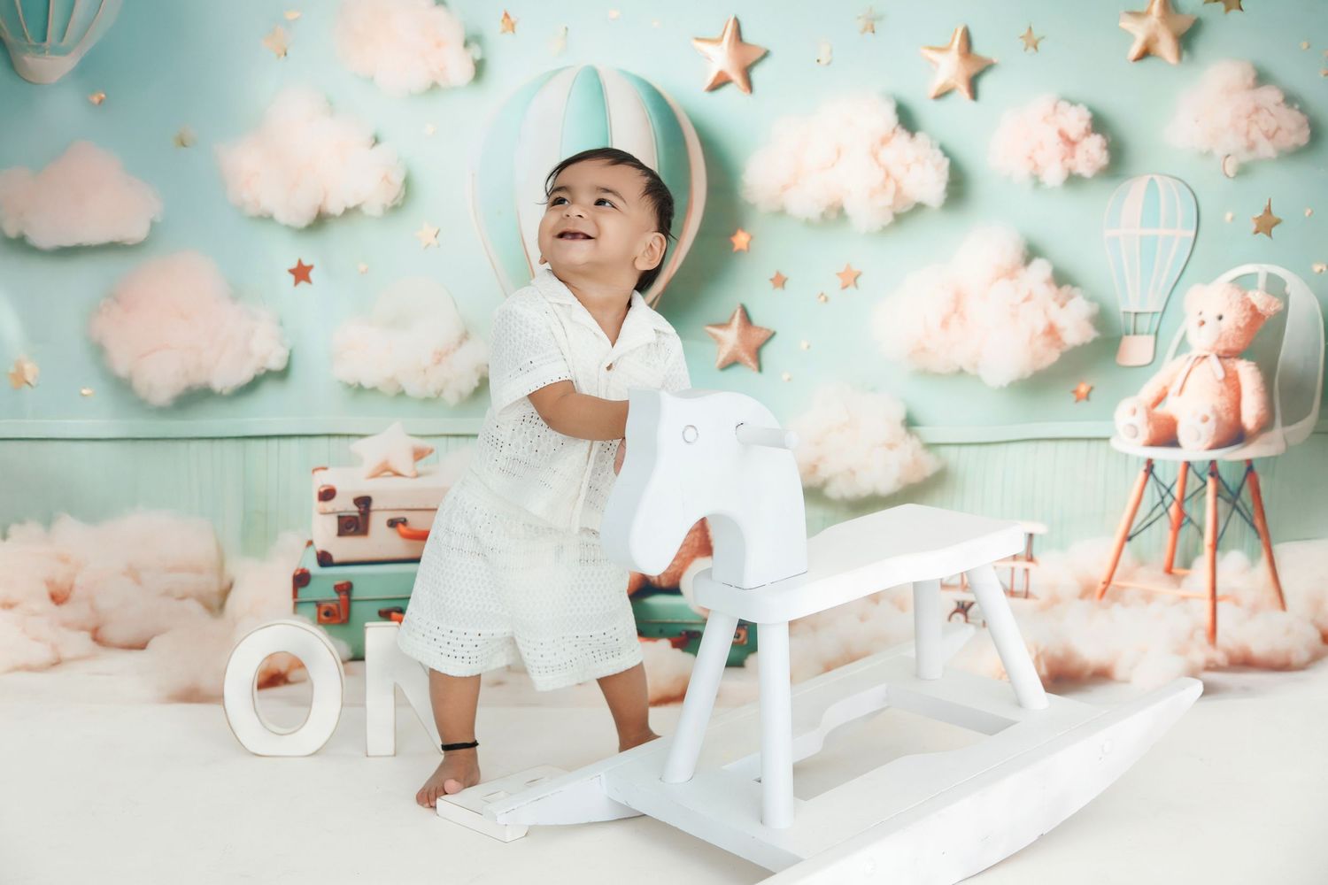 Laughing baby in white outfit riding a white rocking horse against a pastel blue backdrop with clouds and stars.
