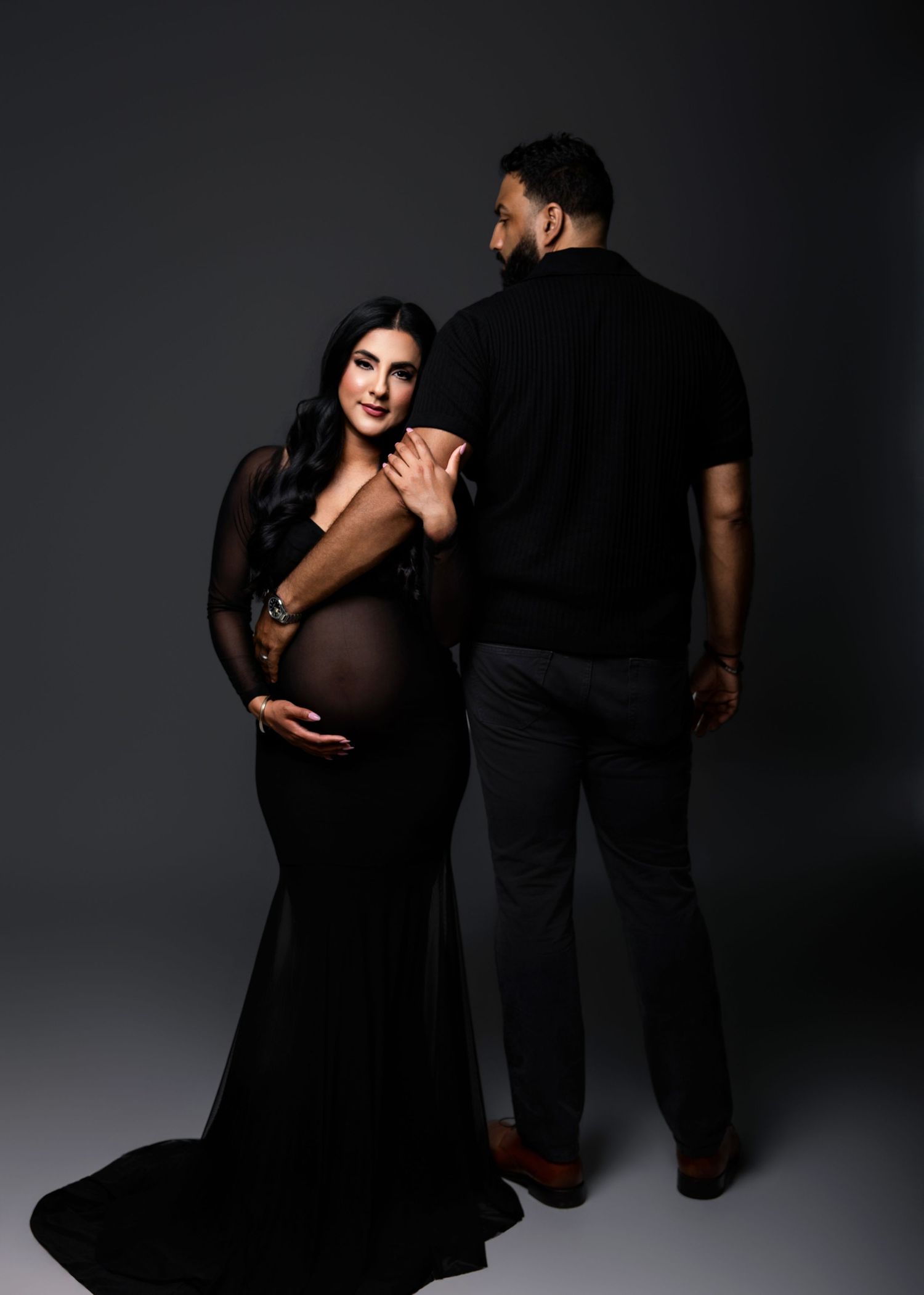 Elegant maternity photo of a pregnant woman in a sheer black gown posing with her partner in a studio.