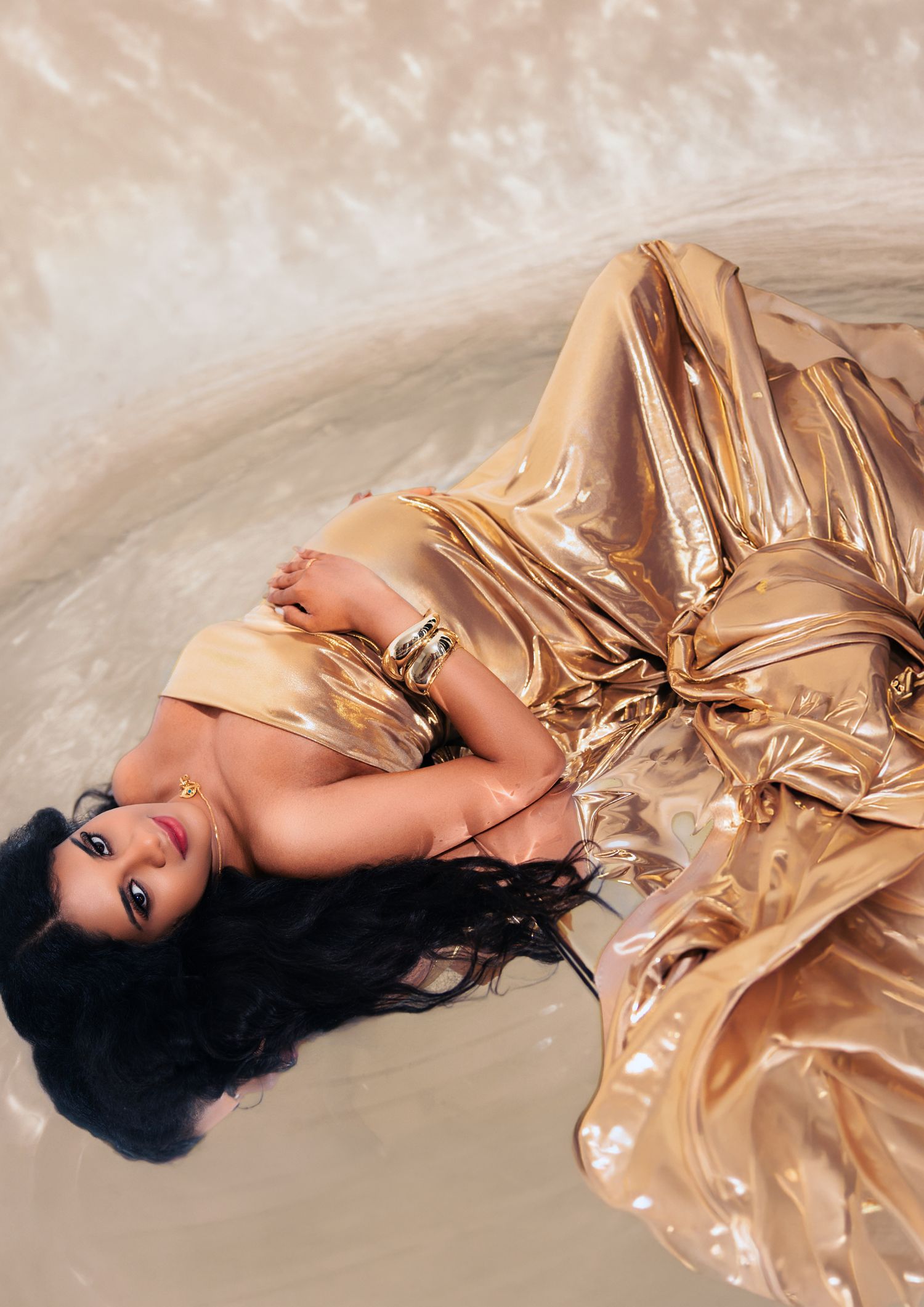 Woman in gold metallic dress lying gracefully in shallow water, wearing gold jewelry with long black hair spread out.