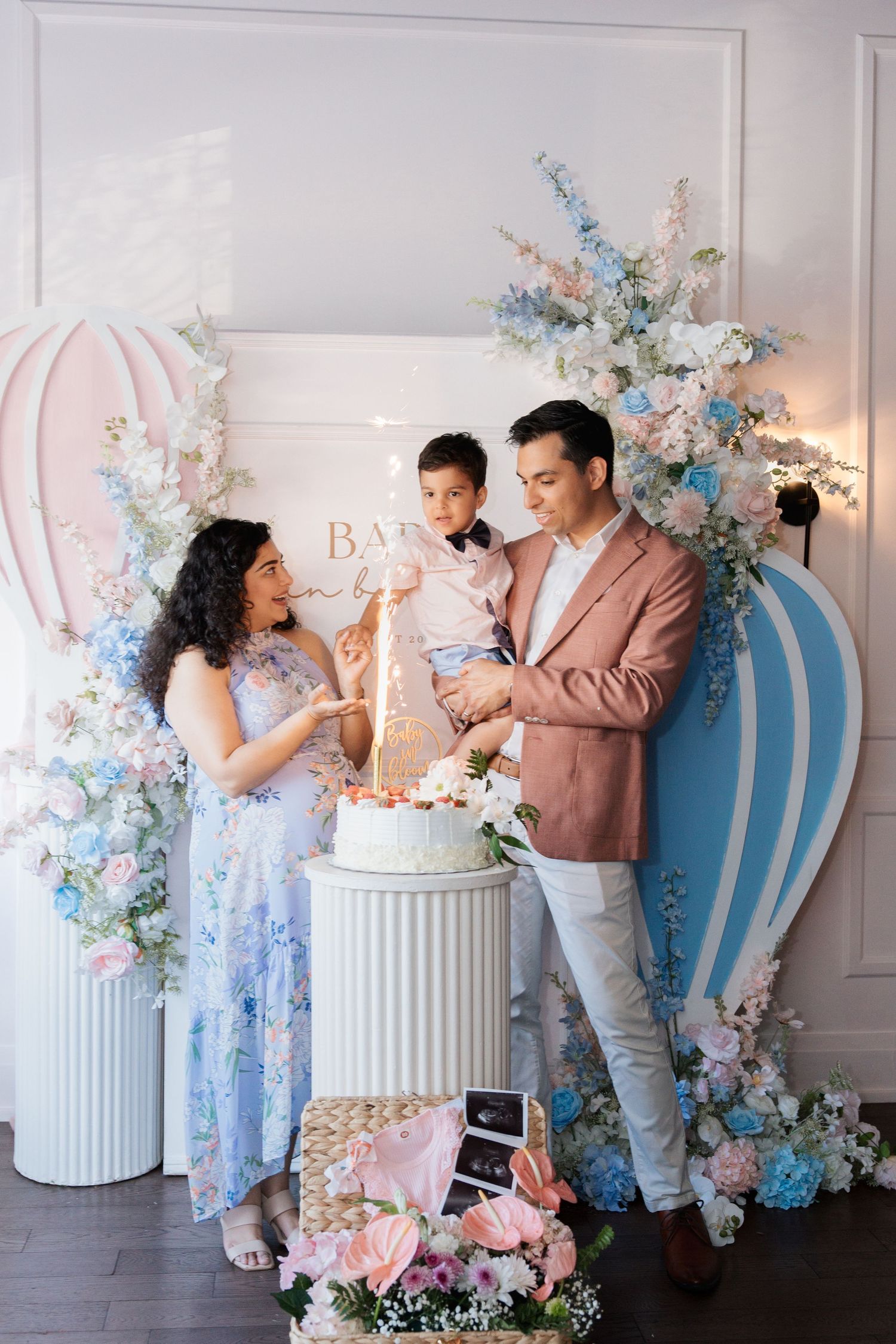 Family gender reveal party with cake, pink and blue floral arch, hot air balloon decor, and ultrasound photos on display.