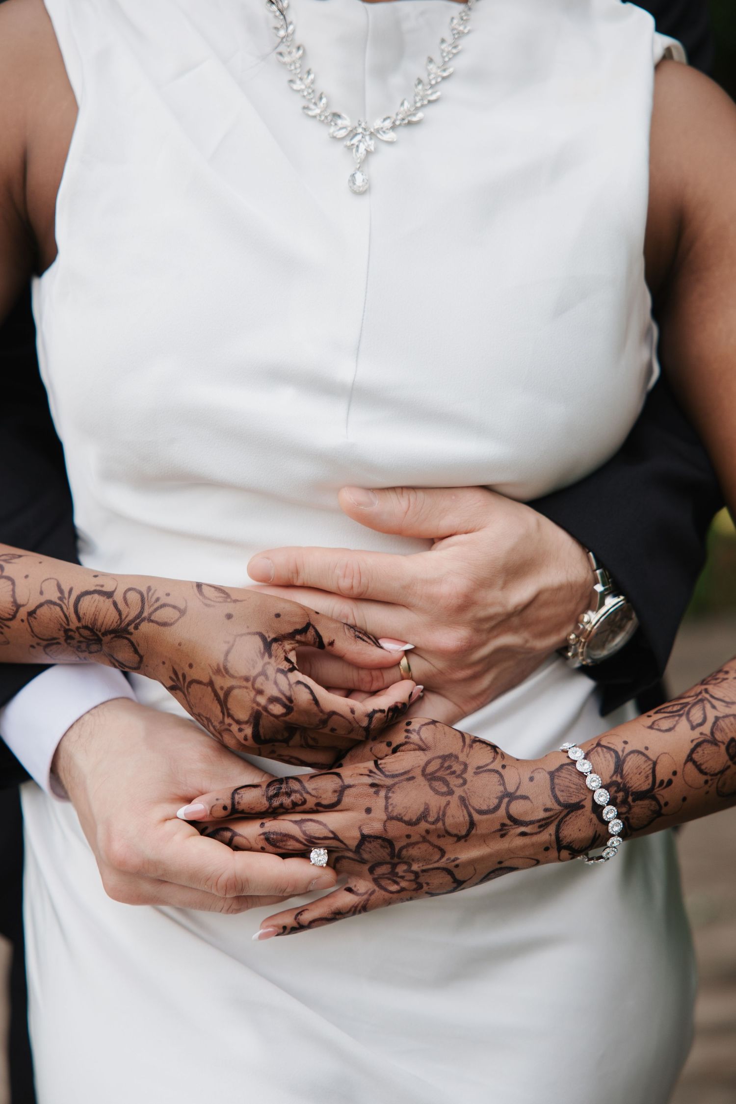 Bride in white dress with henna-adorned hands held by groom, showcasing intricate floral mehndi designs and wedding rings.