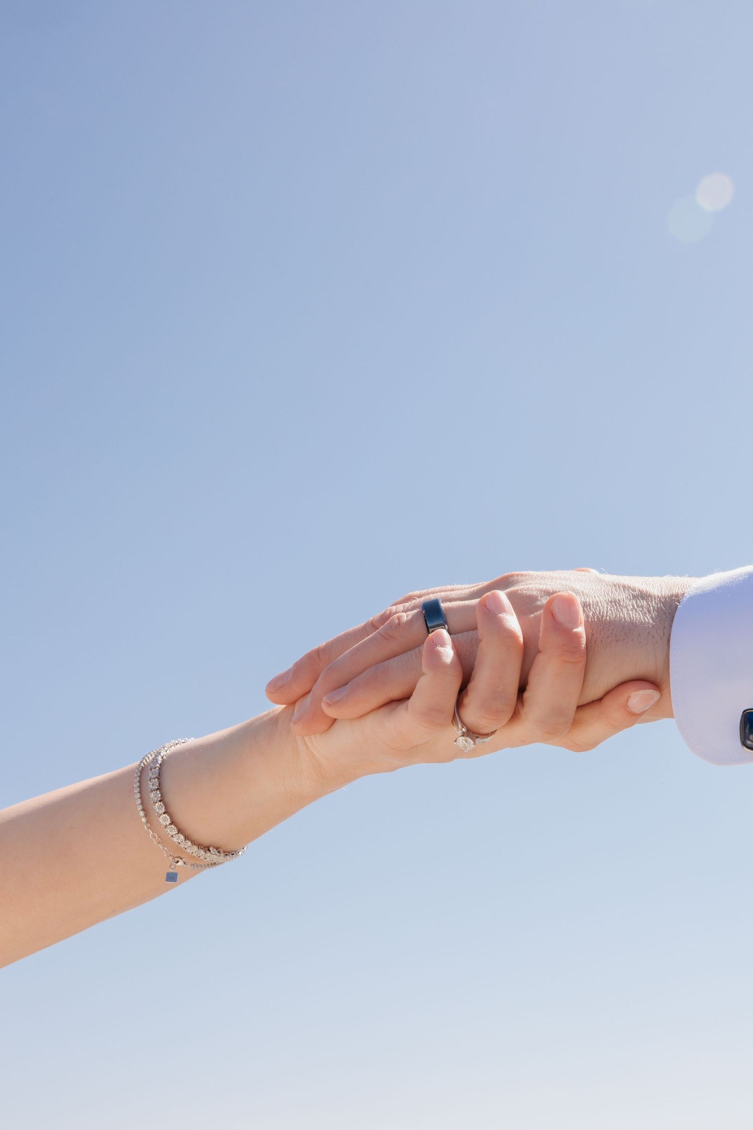 Couple holding hands against blue sky, woman wearing silver bracelet and engagement ring, man in white suit sleeve.