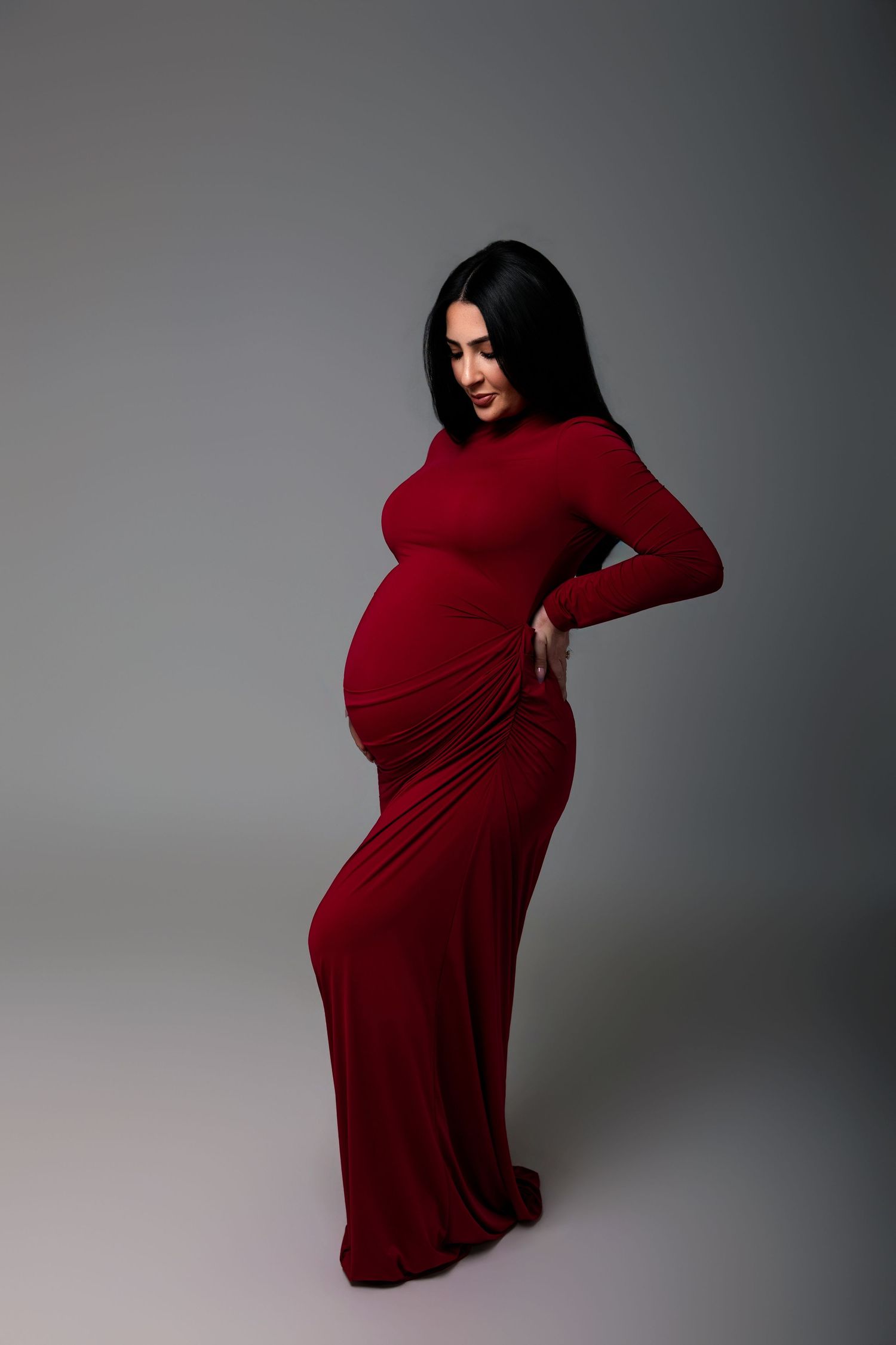 Pregnant woman in elegant red long-sleeve maxi dress posing gracefully in studio, showcasing baby bump.