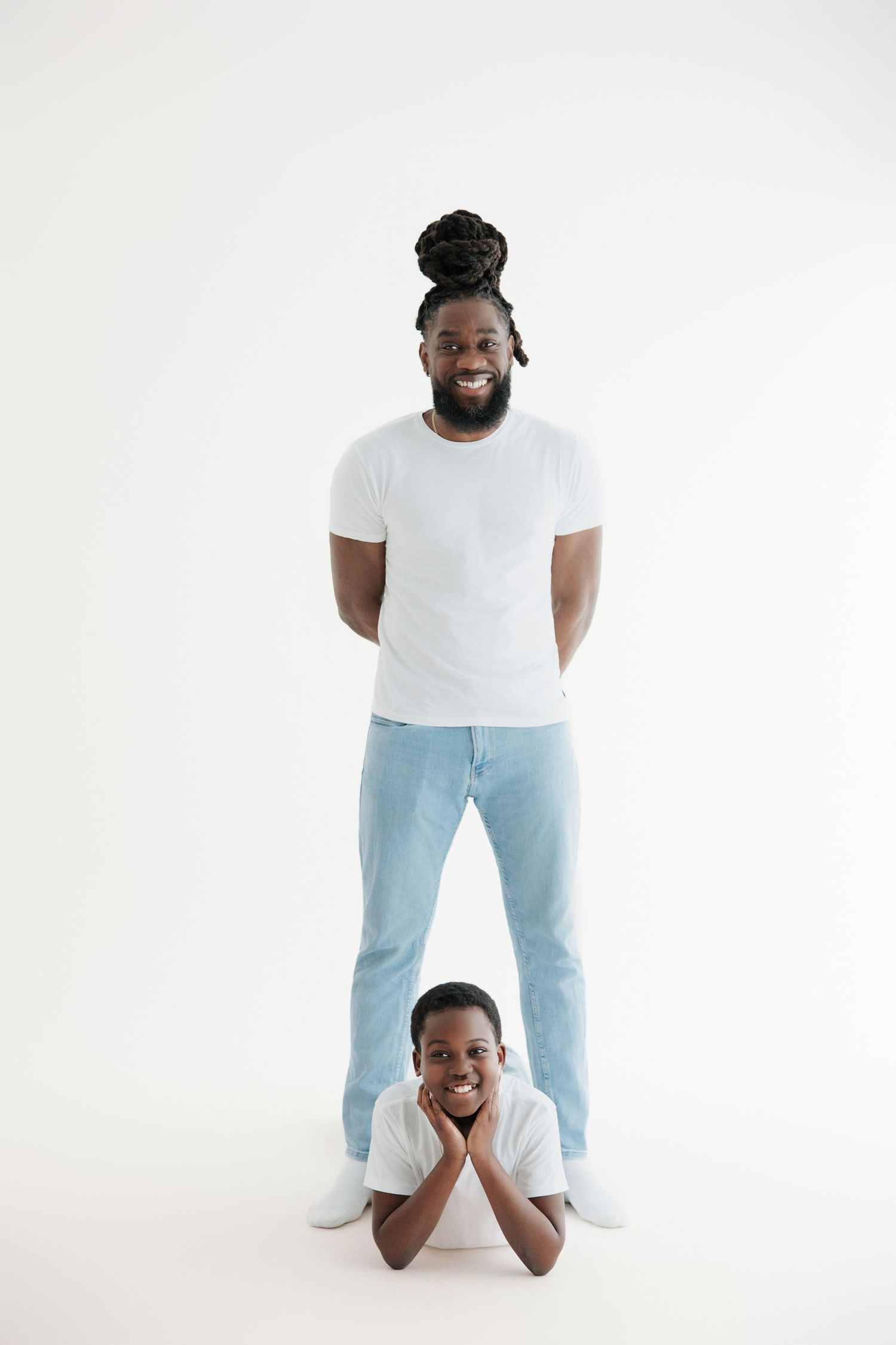 Smiling African American man with dreadlocks stands behind a boy lying on floor, both wearing white tees and jeans.
