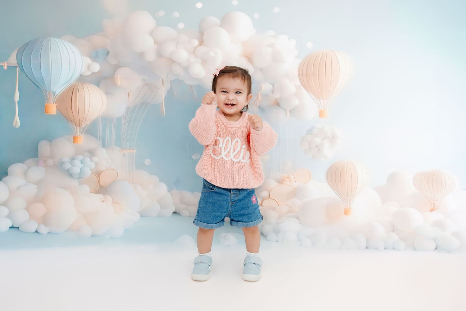 Toddler in pink sweater and denim shorts poses in front of pastel balloon and hot air balloon backdrop.