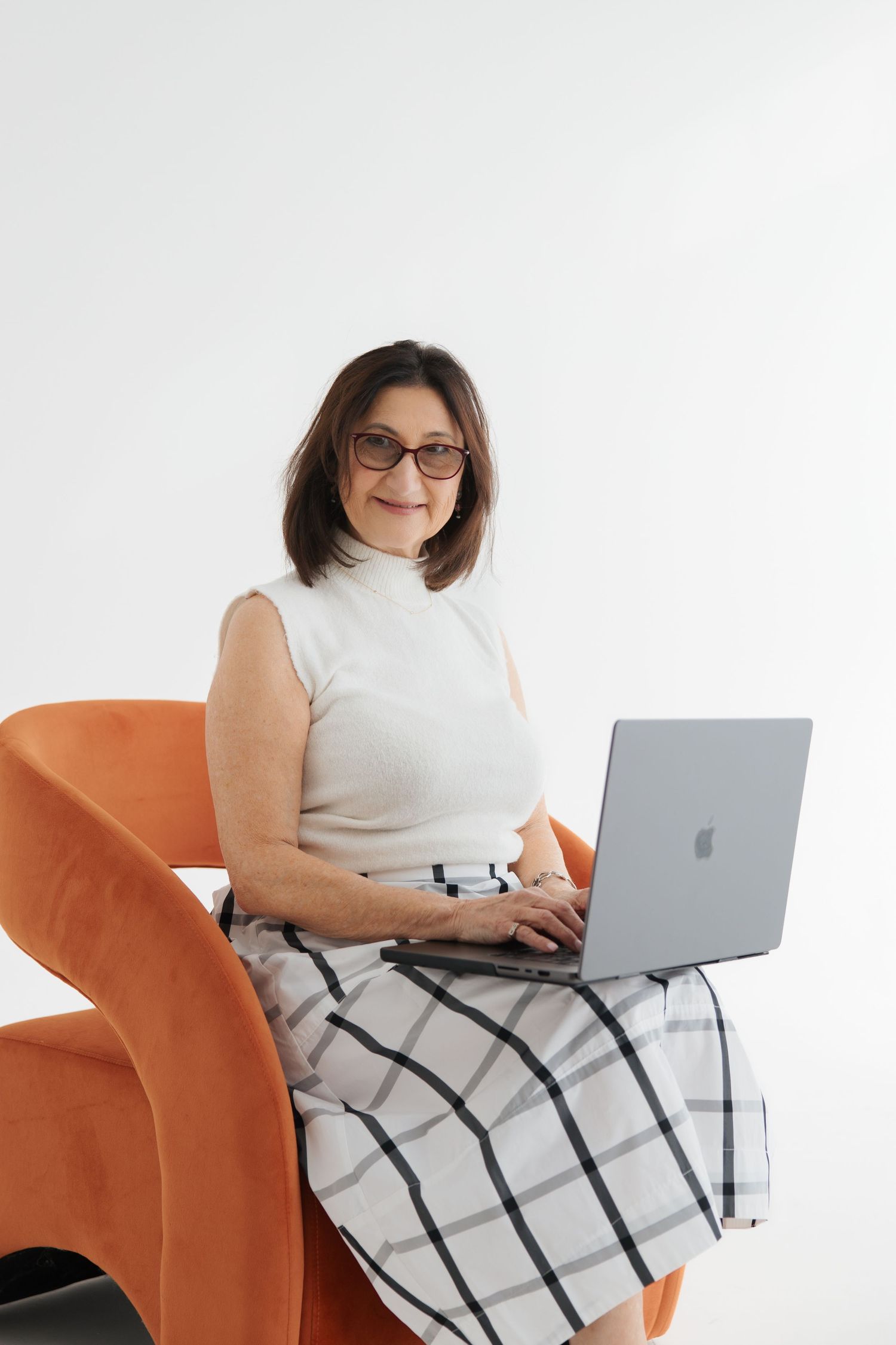 Smiling middle-aged woman with glasses works on a MacBook laptop while seated in an orange chair.