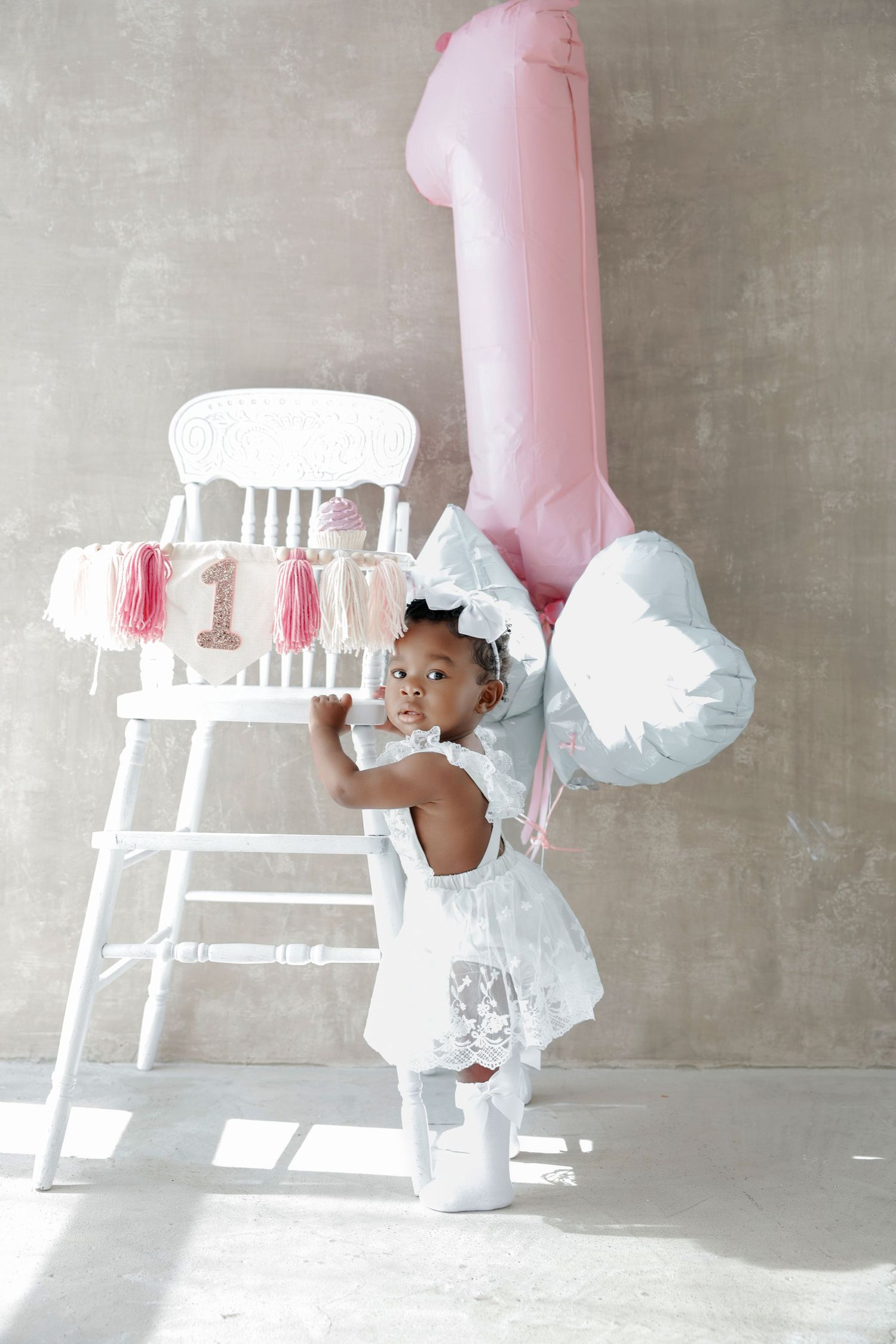 Adorable baby girl in white dress celebrating her 1st birthday with pink balloons and decorated high chair.