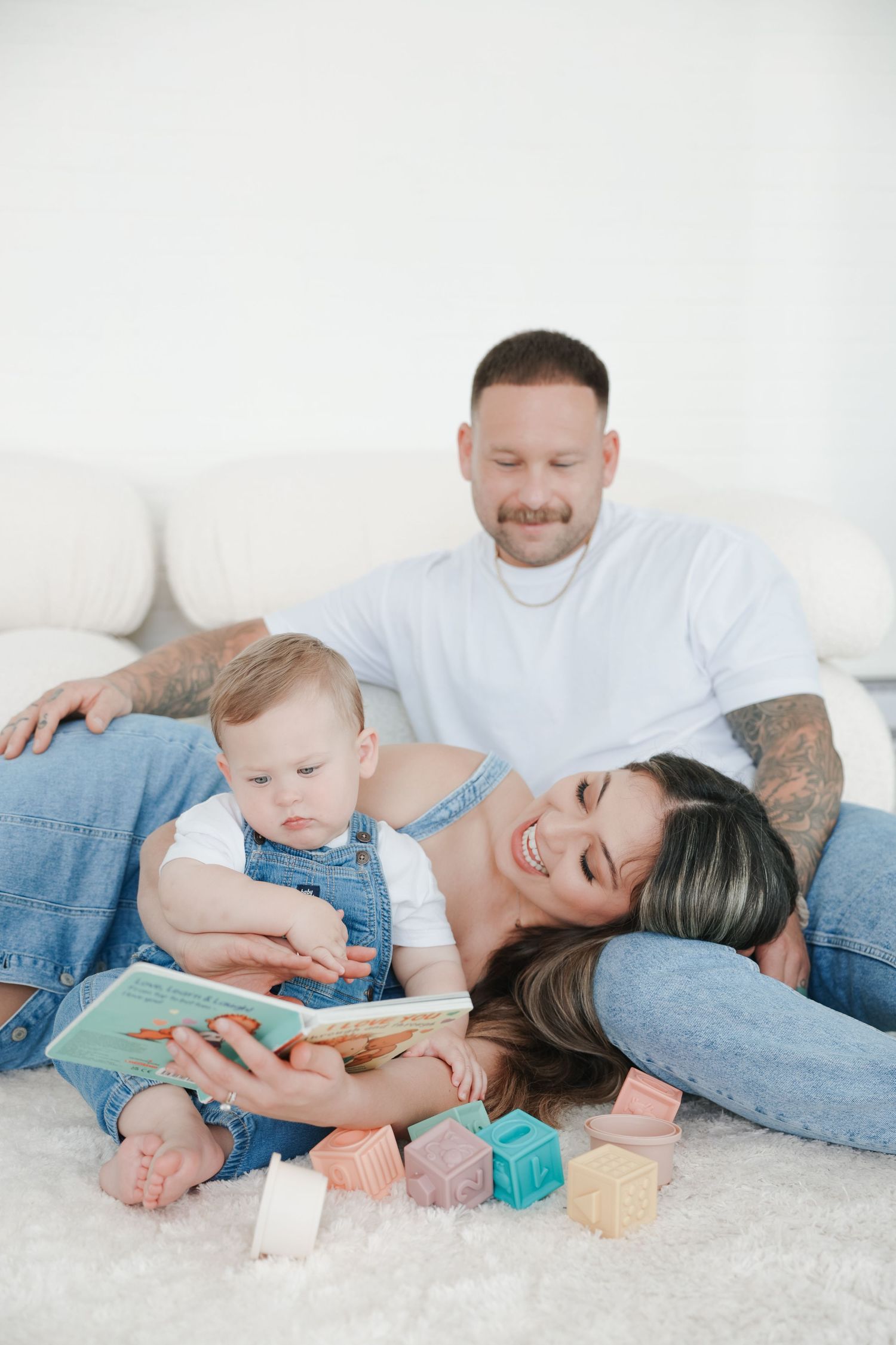 Happy family with tattooed parents and baby reading a colorful book together on a white fluffy carpet at home.