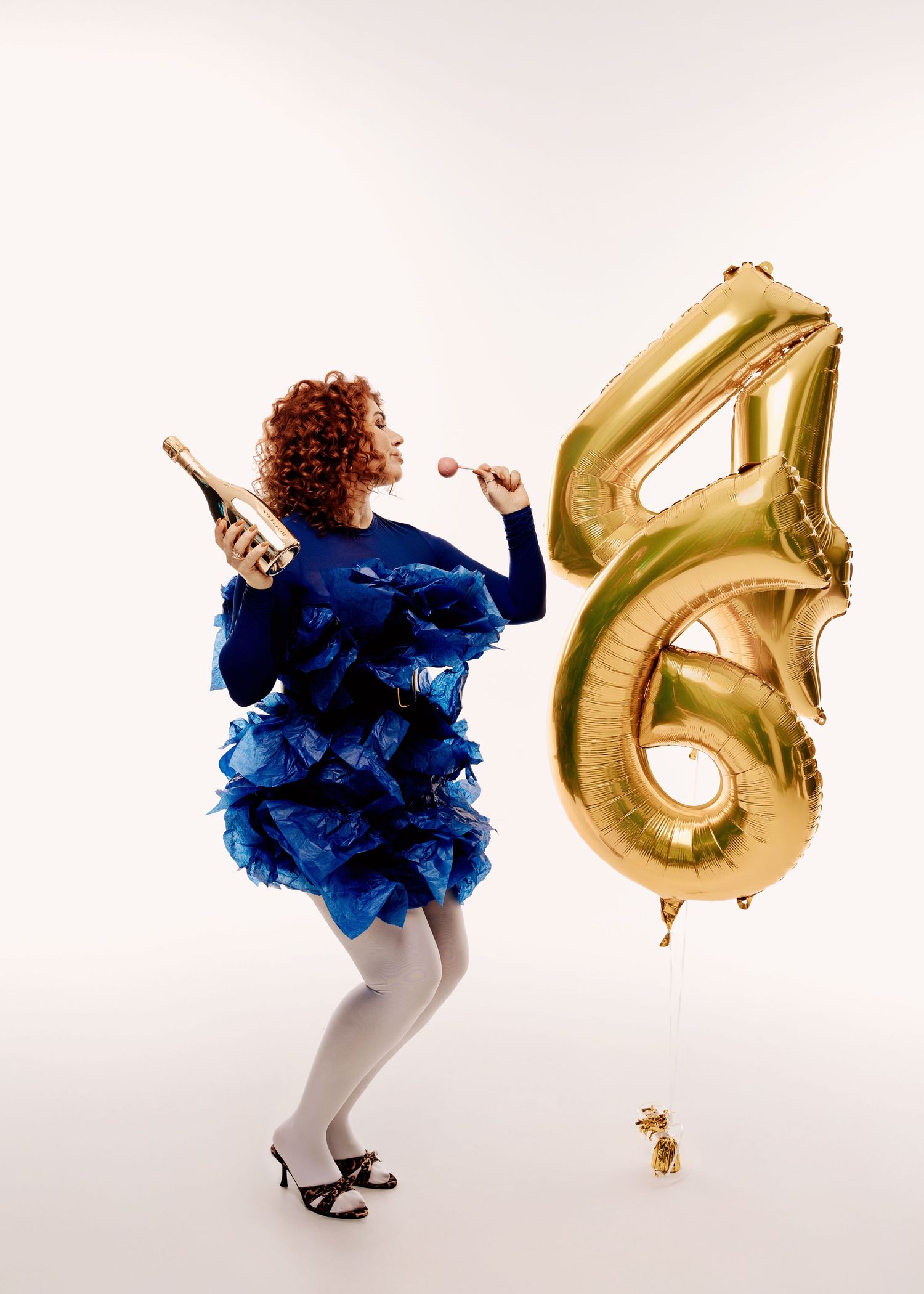Curly redhead woman in a blue ruffled dress holding a champagne bottle beside gold number 46 balloons on white background.