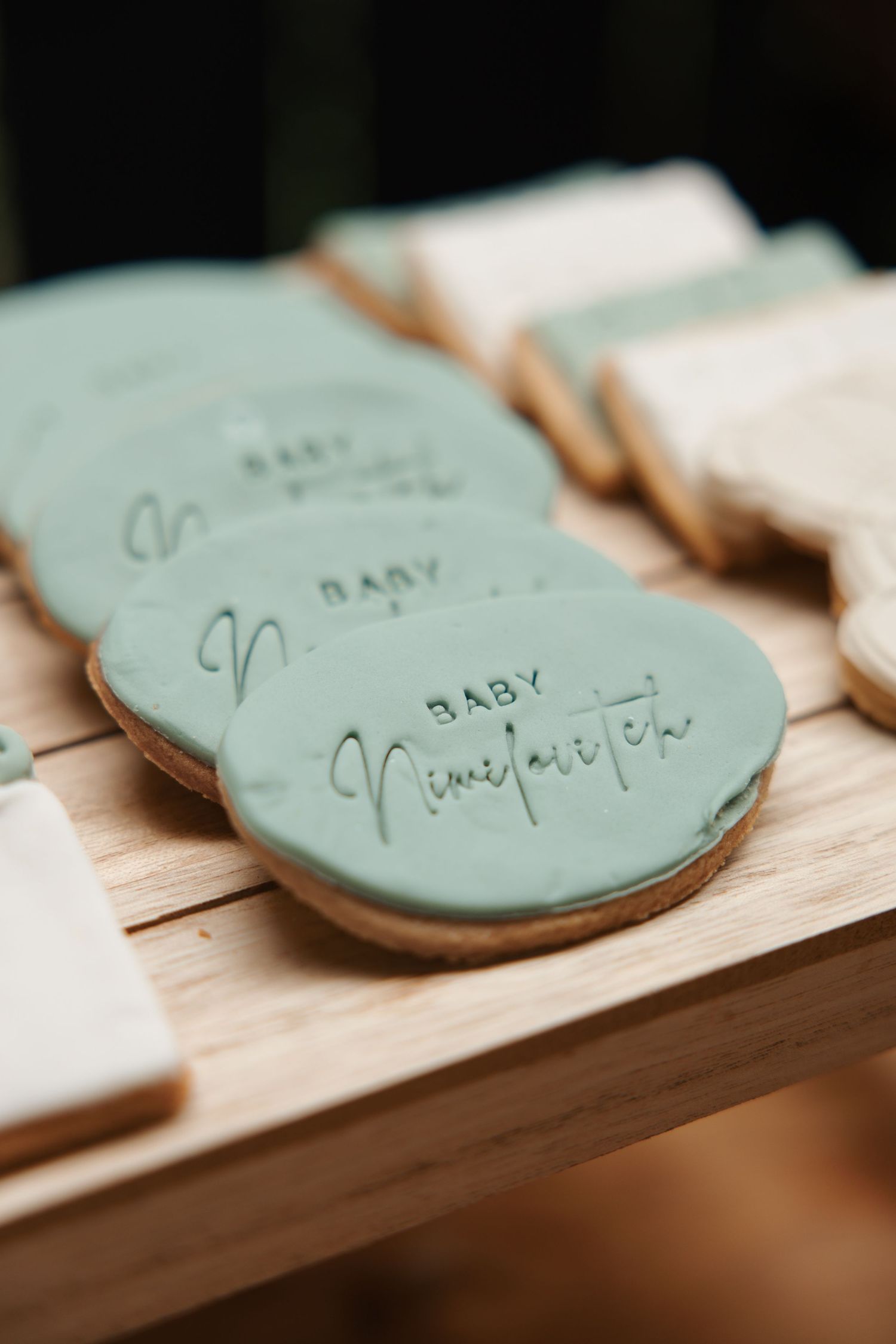 Sage green frosted baby shower cookies with elegant script lettering on a wooden display board.