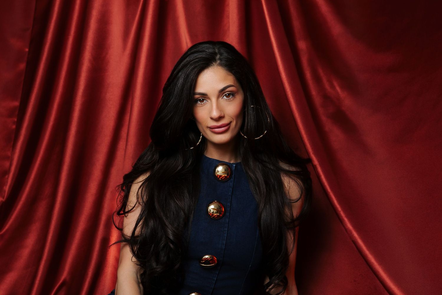 Elegant dark-haired woman in navy top with gold accessories posing before luxurious red velvet curtains.
