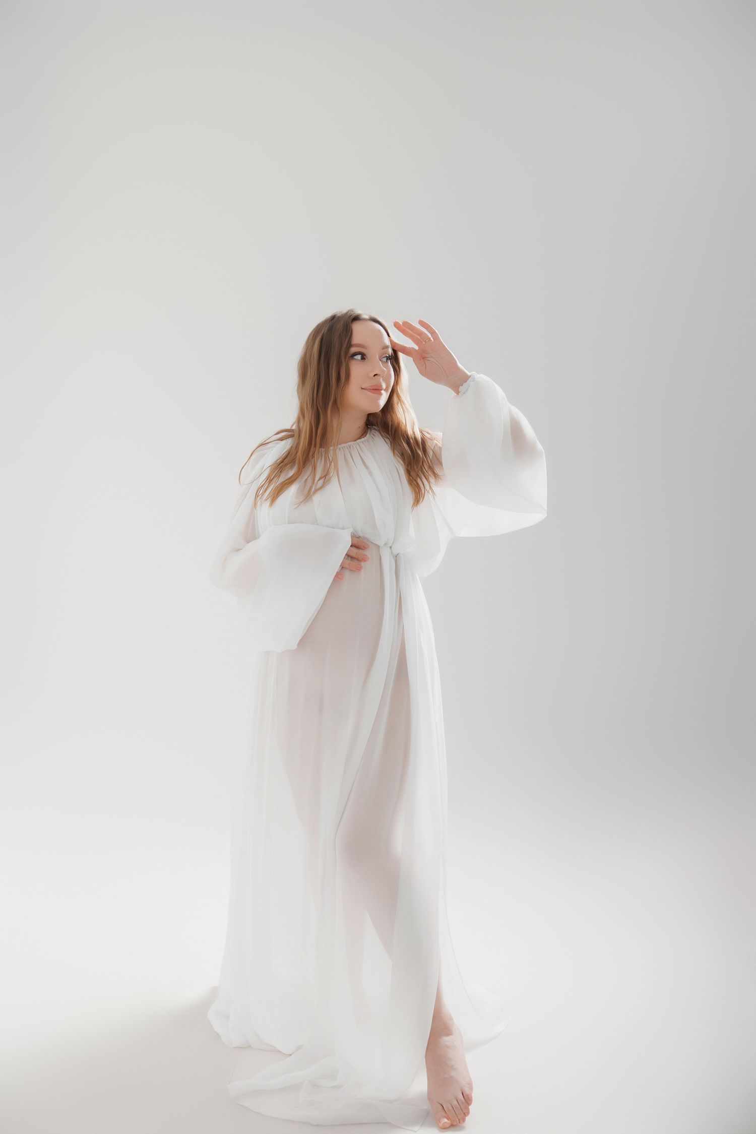 Pregnant woman in flowing white sheer gown poses gracefully against white background in ethereal maternity photo.