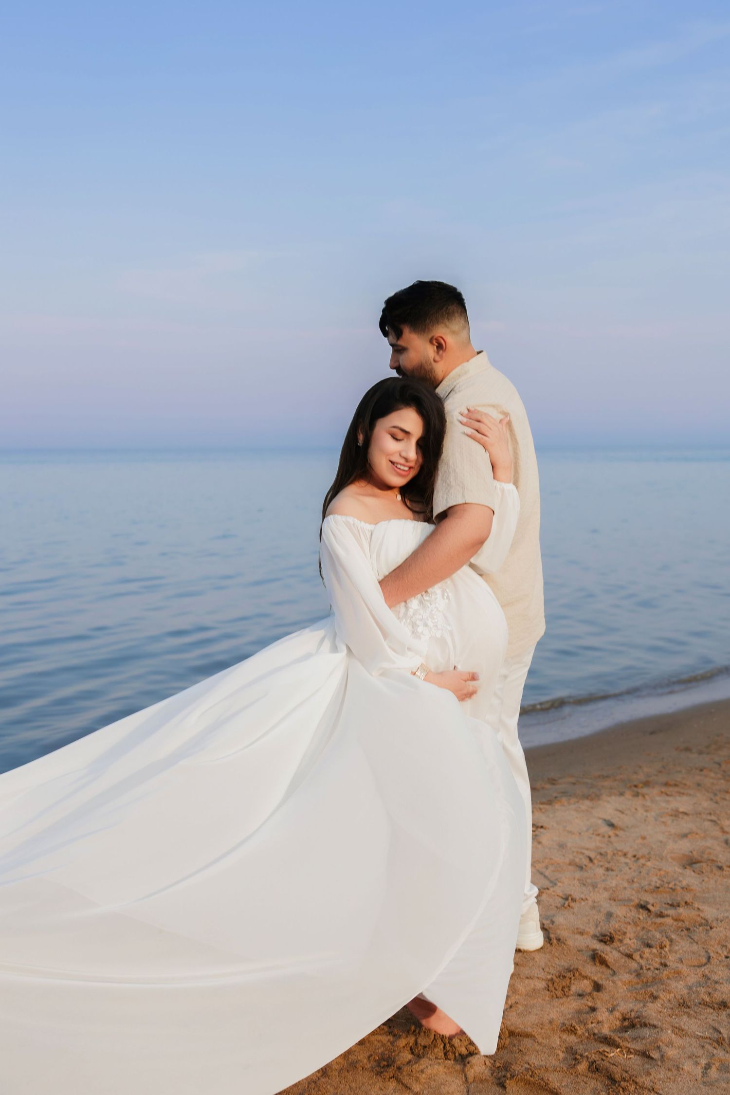Pregnant couple embracing on sandy beach, woman in flowing white dress, man in linen shirt, calm ocean backdrop.
