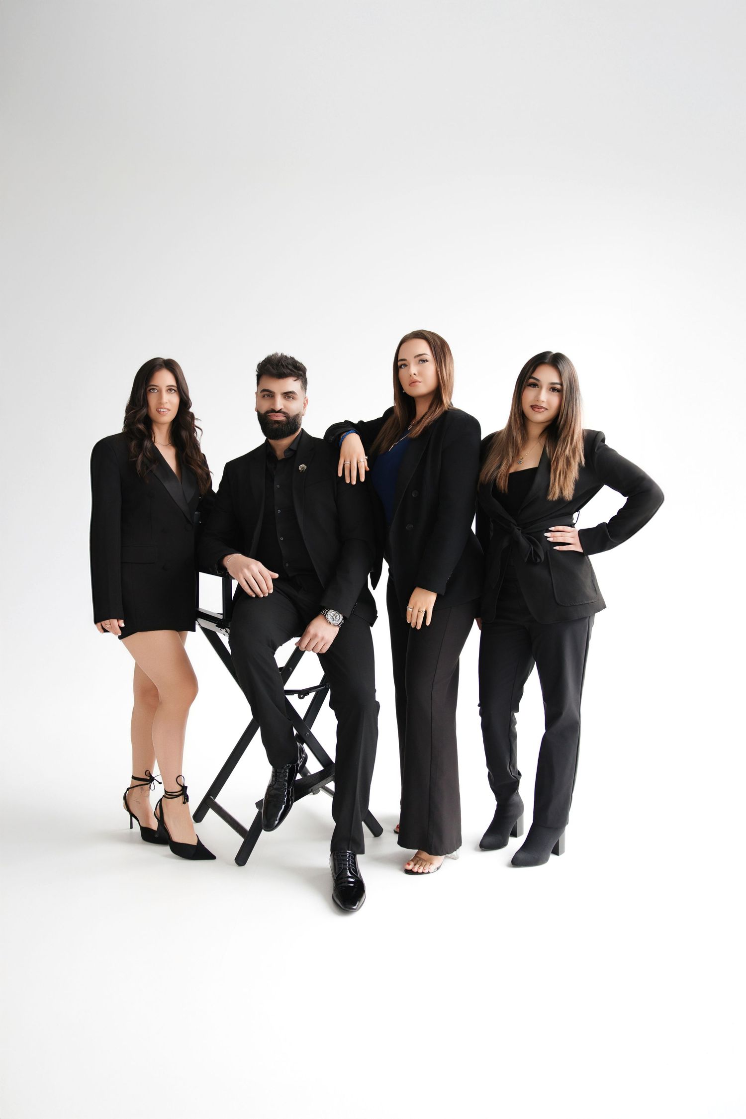 Four professionals in black suits pose confidently in a studio, with one man seated on a director's chair.