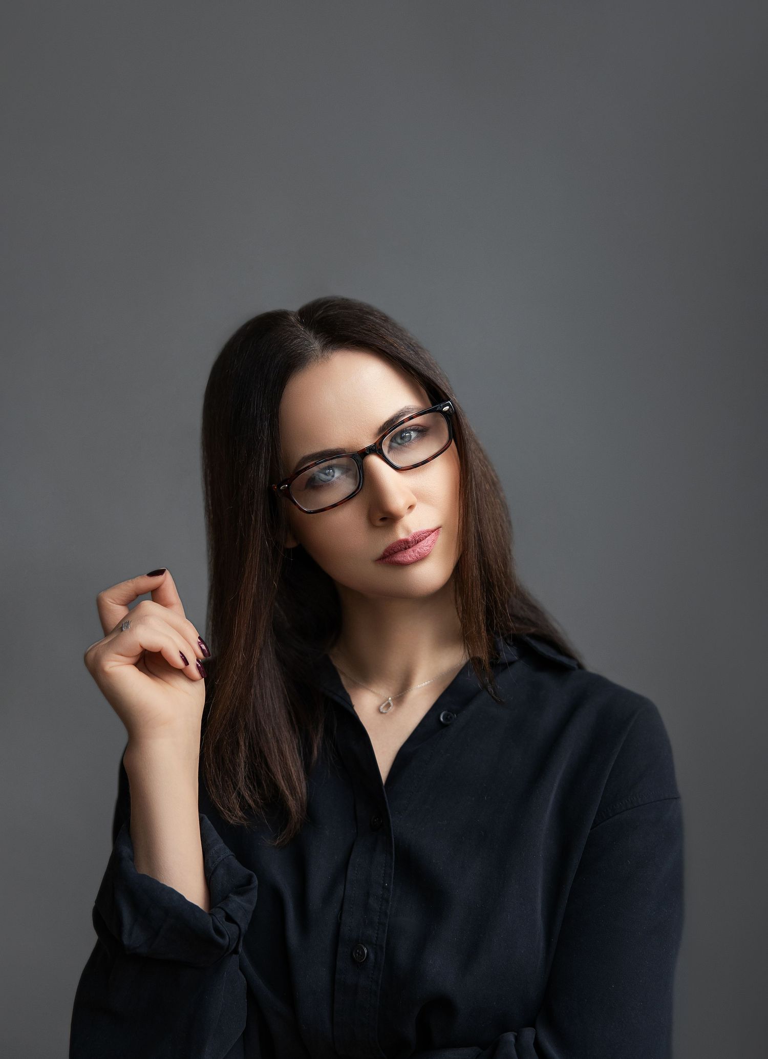 Brunette woman wearing glasses and black shirt posing thoughtfully against gray studio background.