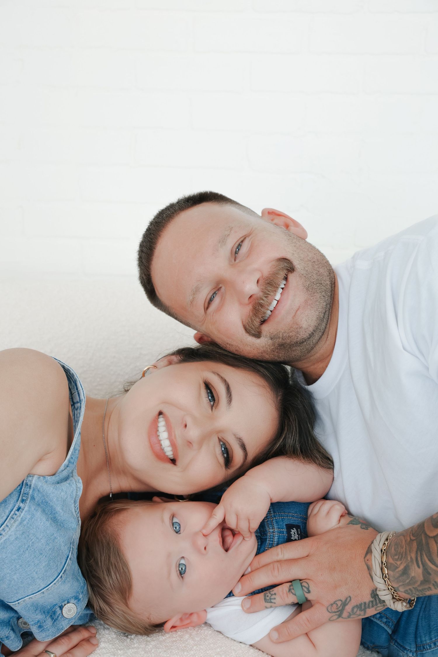 Happy young family with tattooed father, smiling mother, and blue-eyed baby lying together on white background.