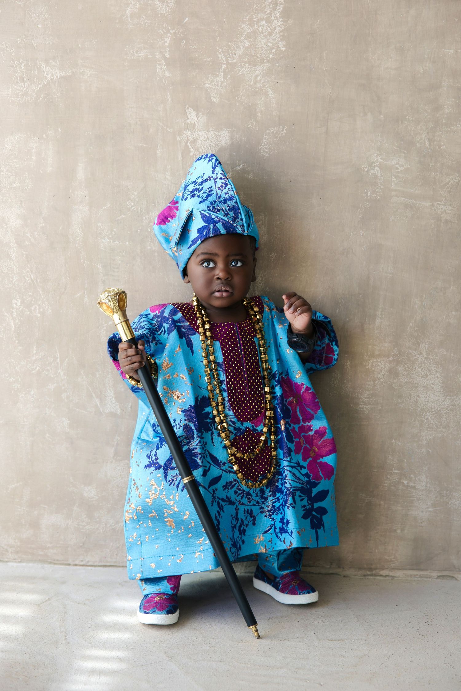 Young child in vibrant blue African traditional attire with gold beaded necklaces and ornate staff, posing confidently.