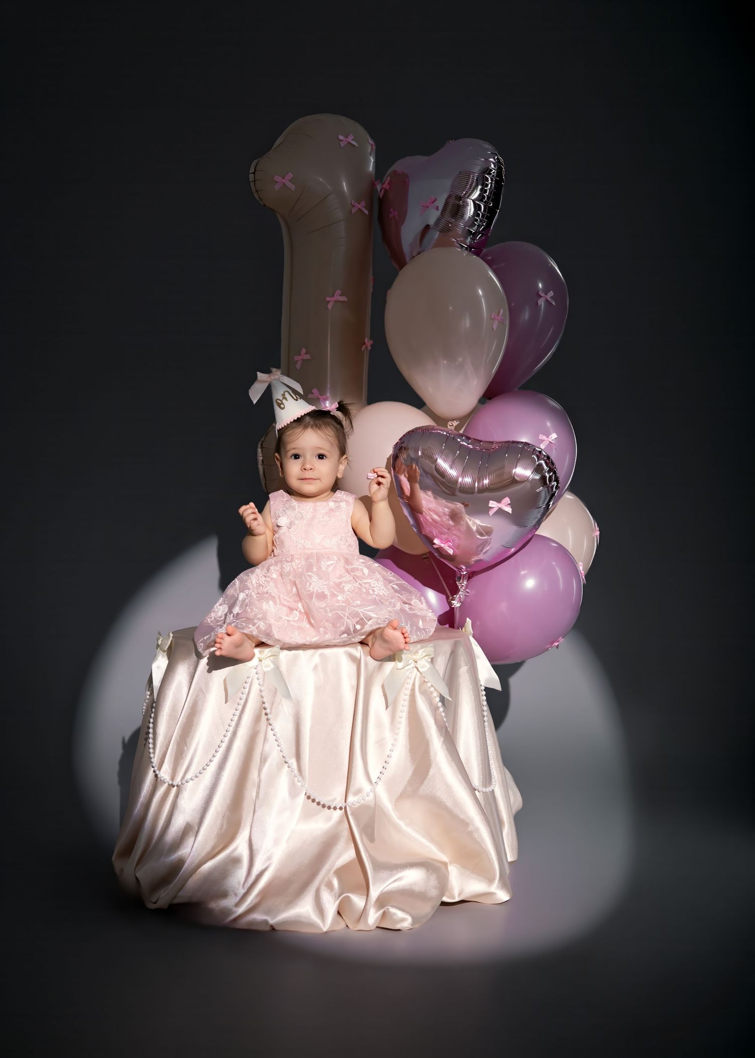 Baby girl in pink dress celebrates first birthday with number one balloon and pink heart balloons in studio photo.