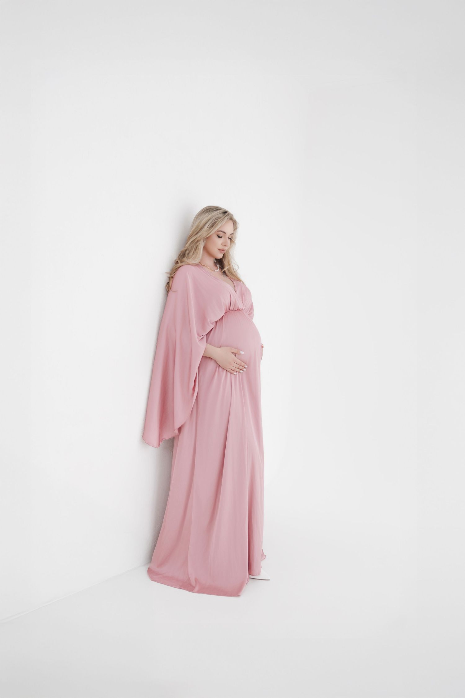 Pregnant woman in blush pink maxi dress with flowing sleeves, cradling baby bump against white background.